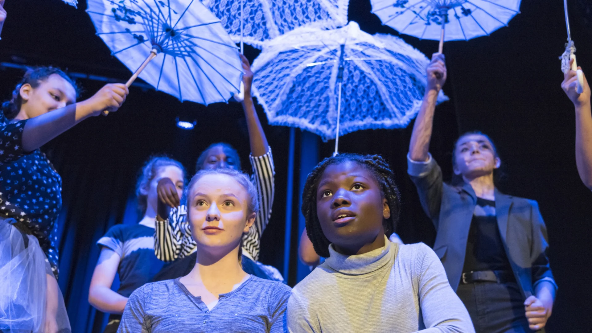 A photo of two young girls knelt down at the front of the stage with white umbrellas held over their heads.