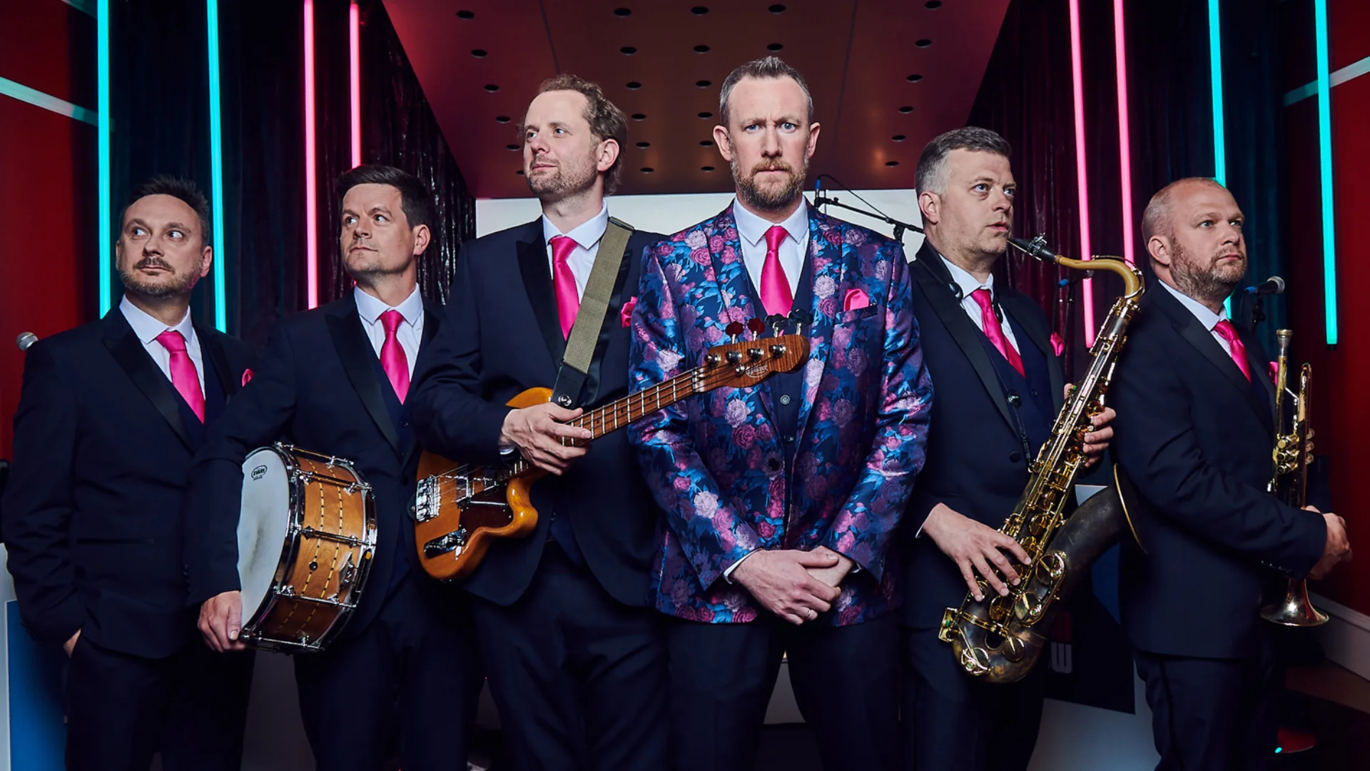 A group of musicians dressed in suits with bright pink ties stand in a line holding their instruments.