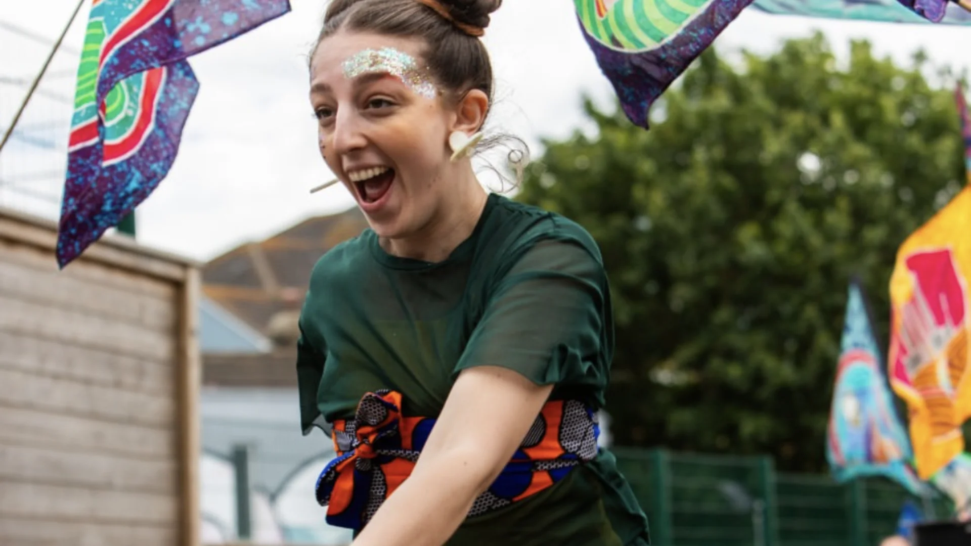 A photo of a white woman with brown hair in two buns, wearing a green t-shirt and glittery face make up. She is outside in a garden surrounded by two flags and smiling.