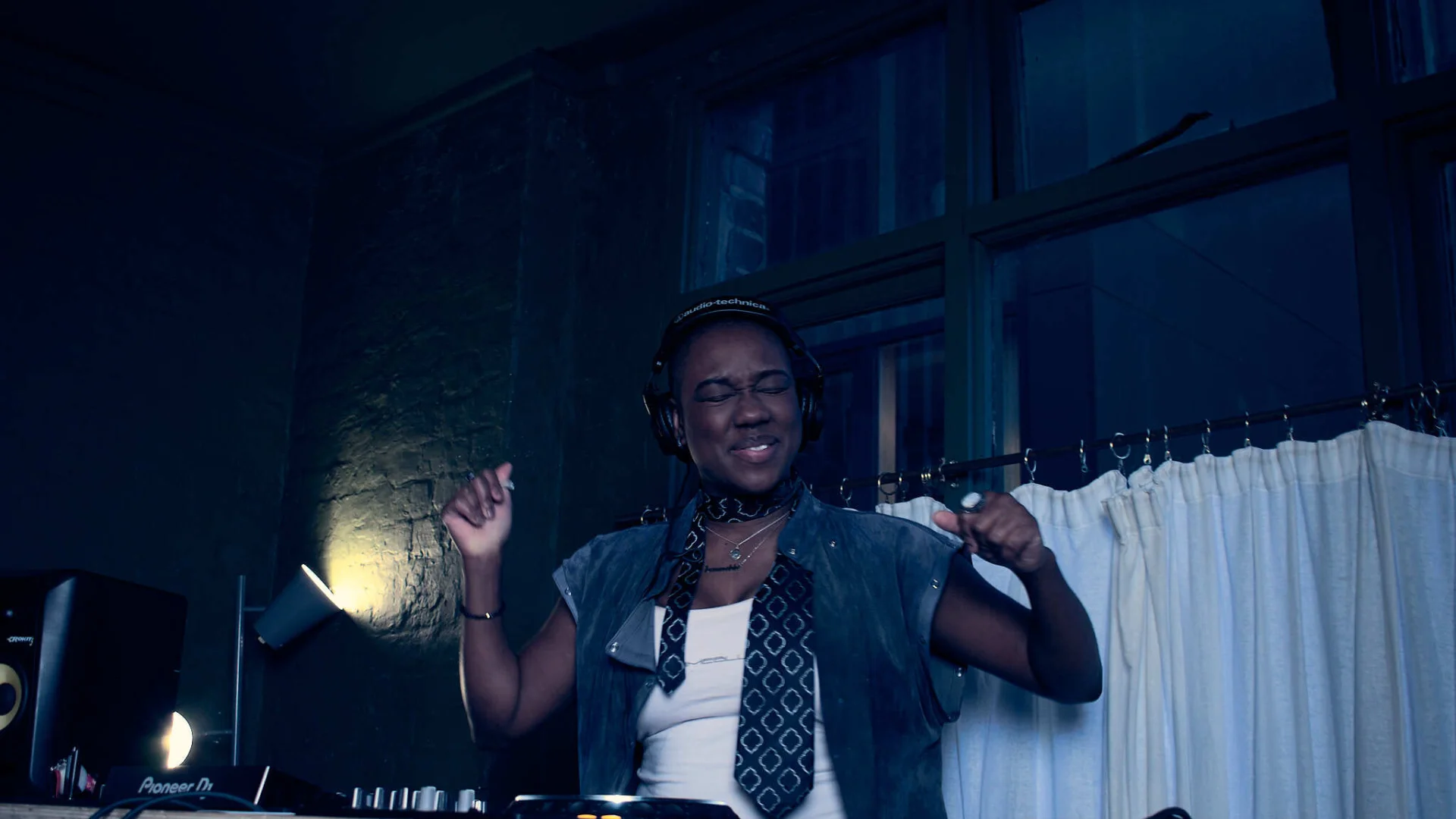 A photo of a black woman stood with her hands in the air behind a set of DJ decks.