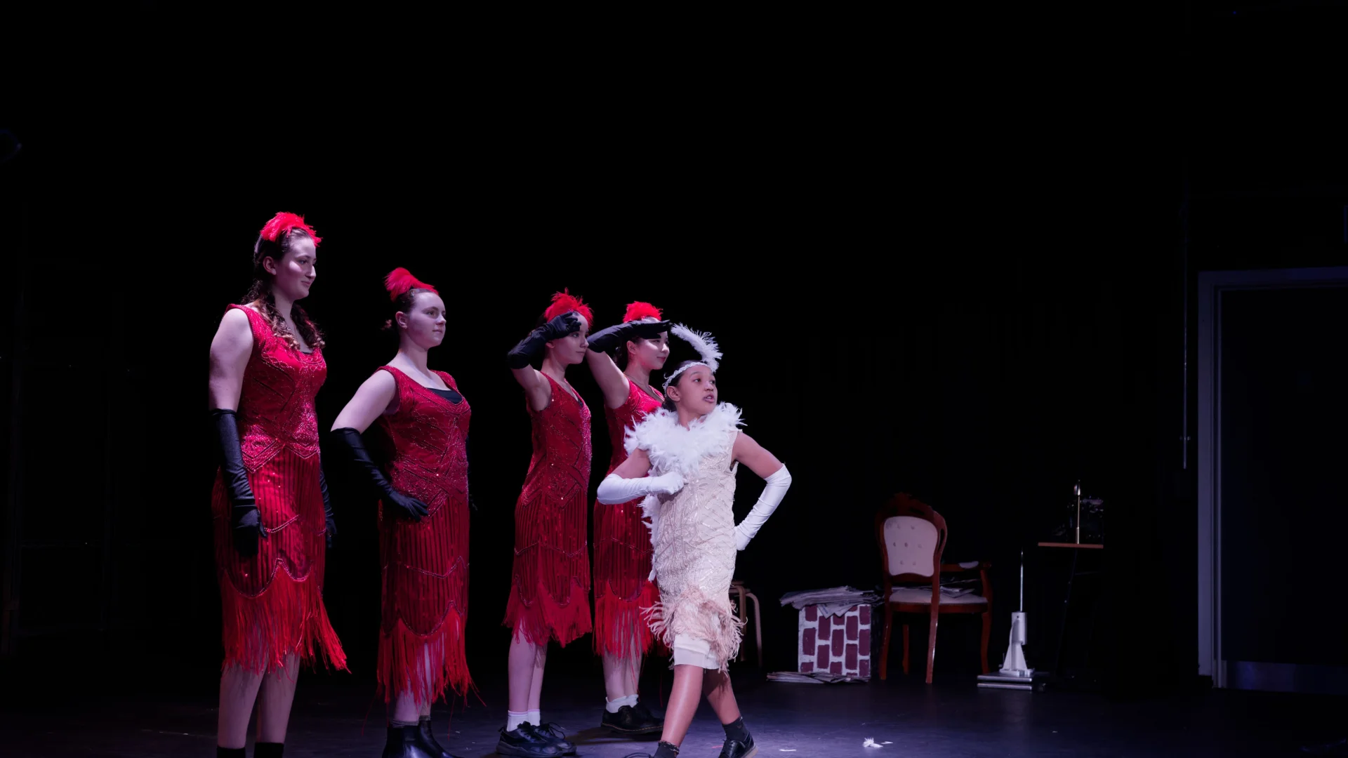 A photo of a group of young performers on stage. They are dressed in 1920s dresses and feather hats. There is a line of performers in red dresses and one in white struts in front of them.
