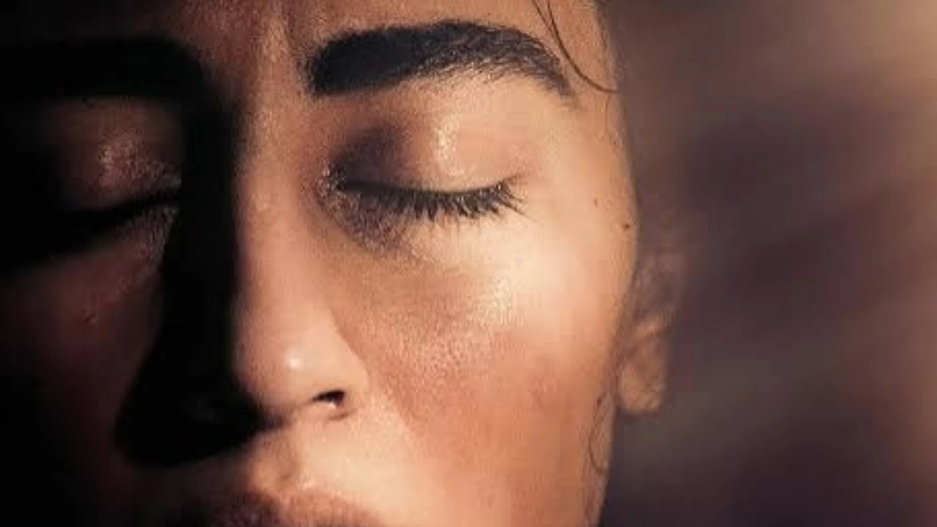 A closeup photo of a presumed woman's face with eyes closed, her left side is in darkness, but light pours in from the top right corner, juxtaposing light against the shadows of what lies in the darkness.