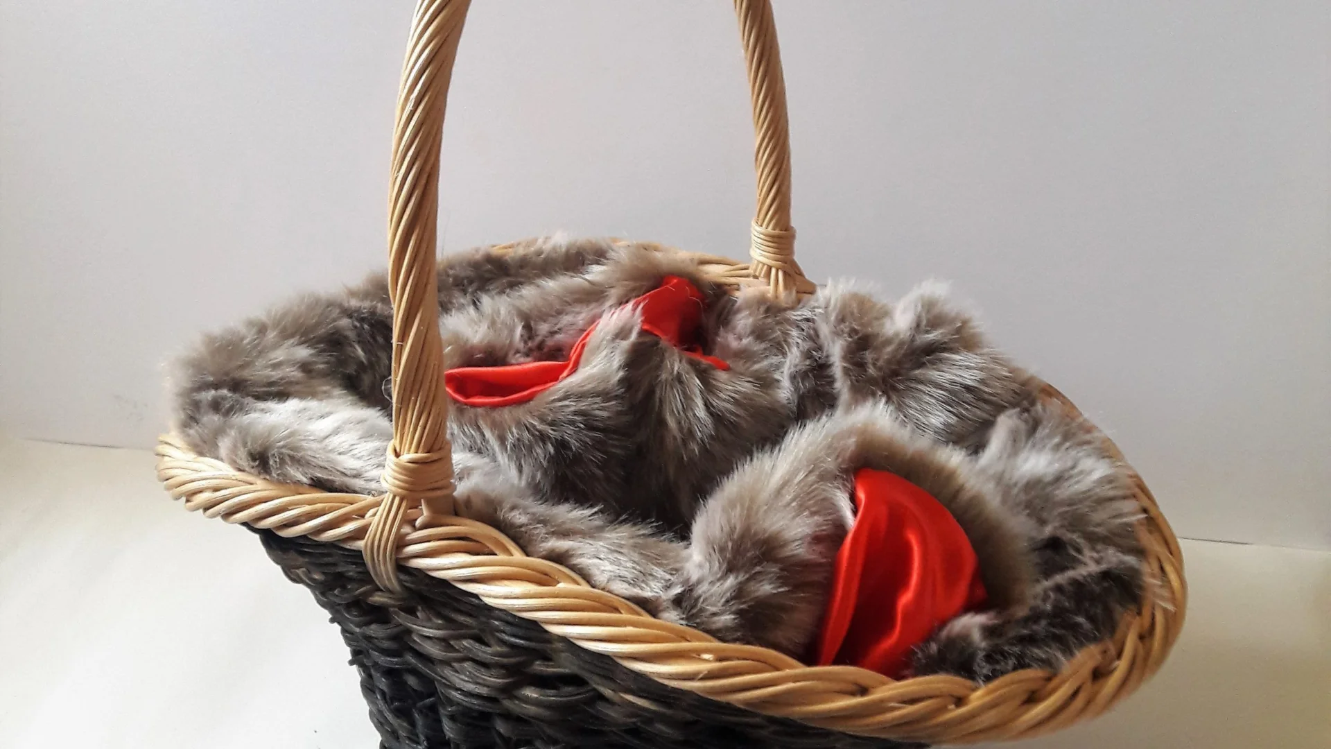 A woven with a darker material body and a light wicker handle is filled with grey fur lined with a red fabric. The fur and fabric mix is mixed in the basket so it all seems to be a part of the same item. The basket is set on a white background.