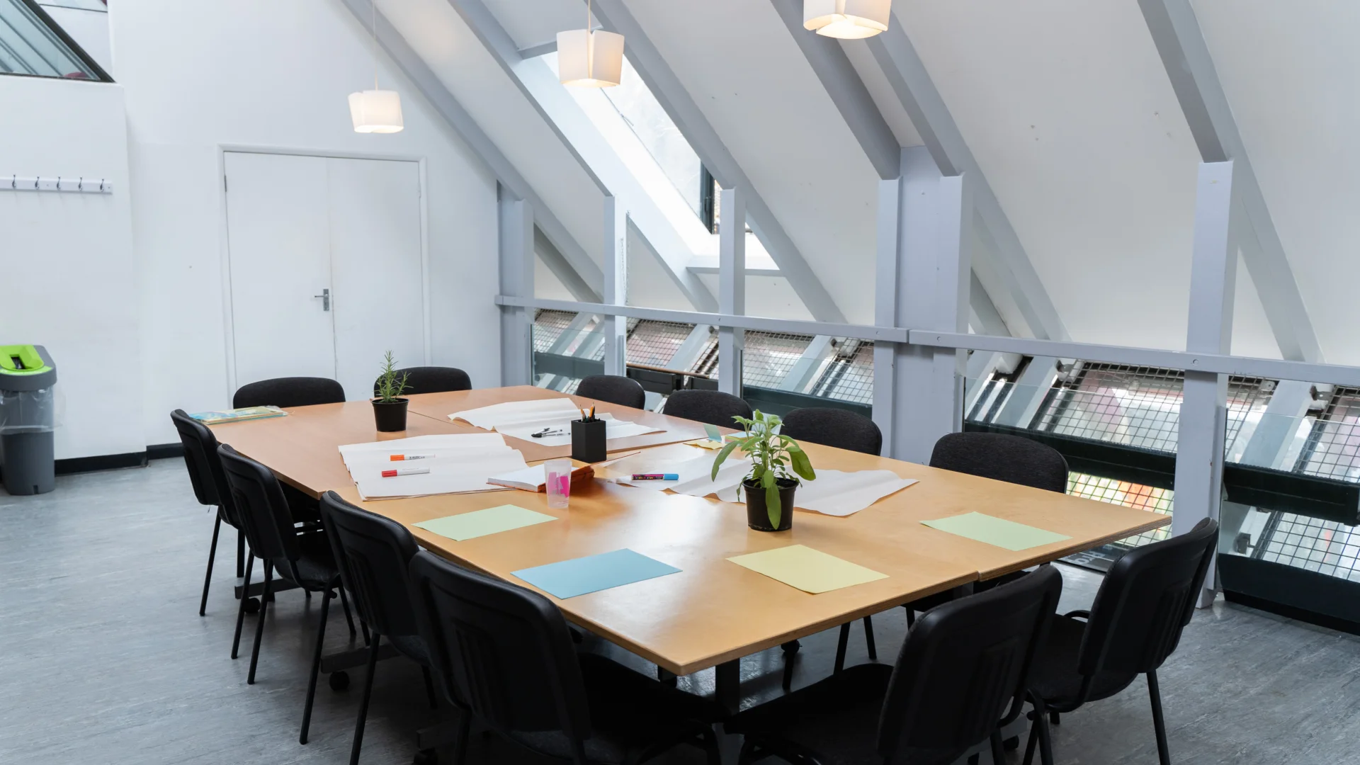 A photo of a meeting room with an empty table and chairs in the middle, set for a meeting with papers, pens and pot plants.