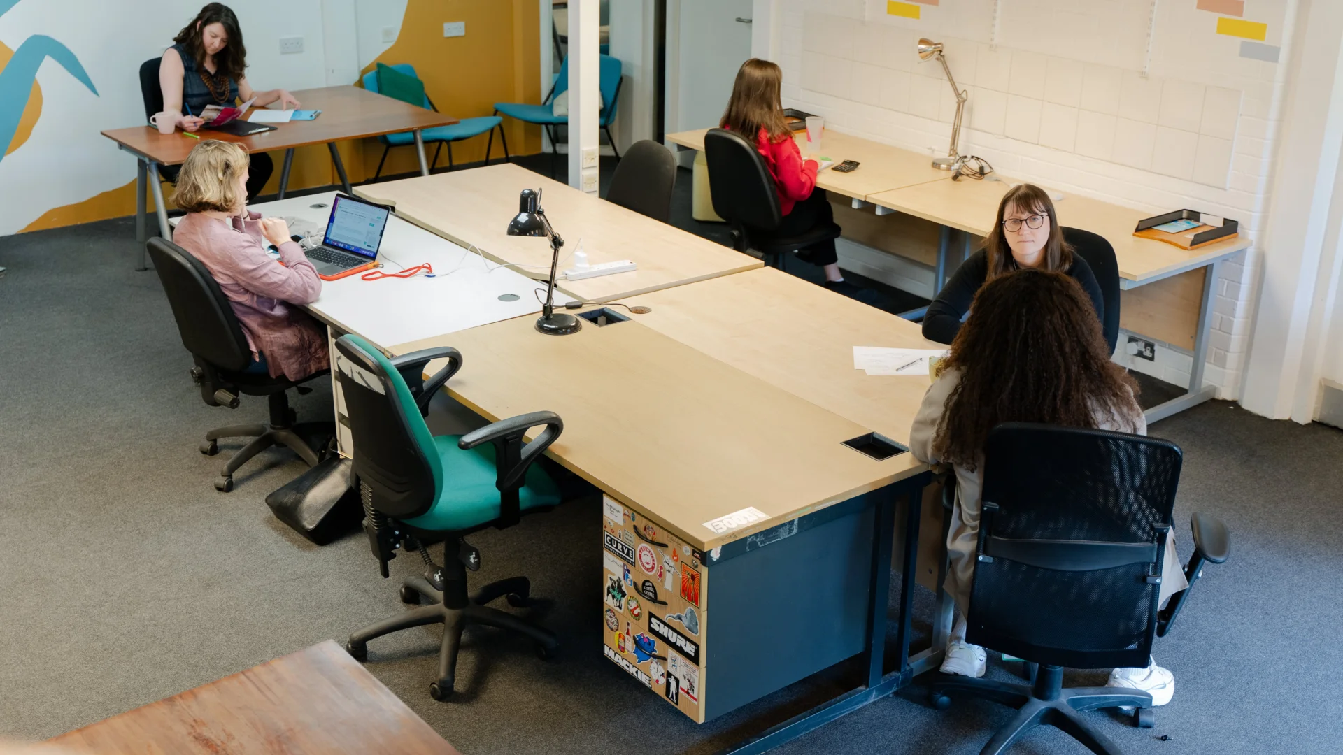 A photo of the Albany coworking space from above. Five people are sat working around a central table and two desks. The walls are covered with colourful murals.