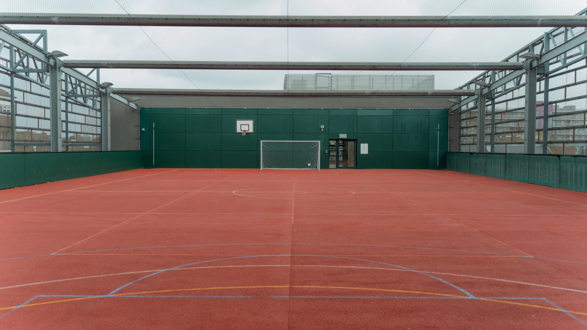 A photo of a ball court with red floor and green walls. The court is open to the elements and it is a grey, cloudy day.