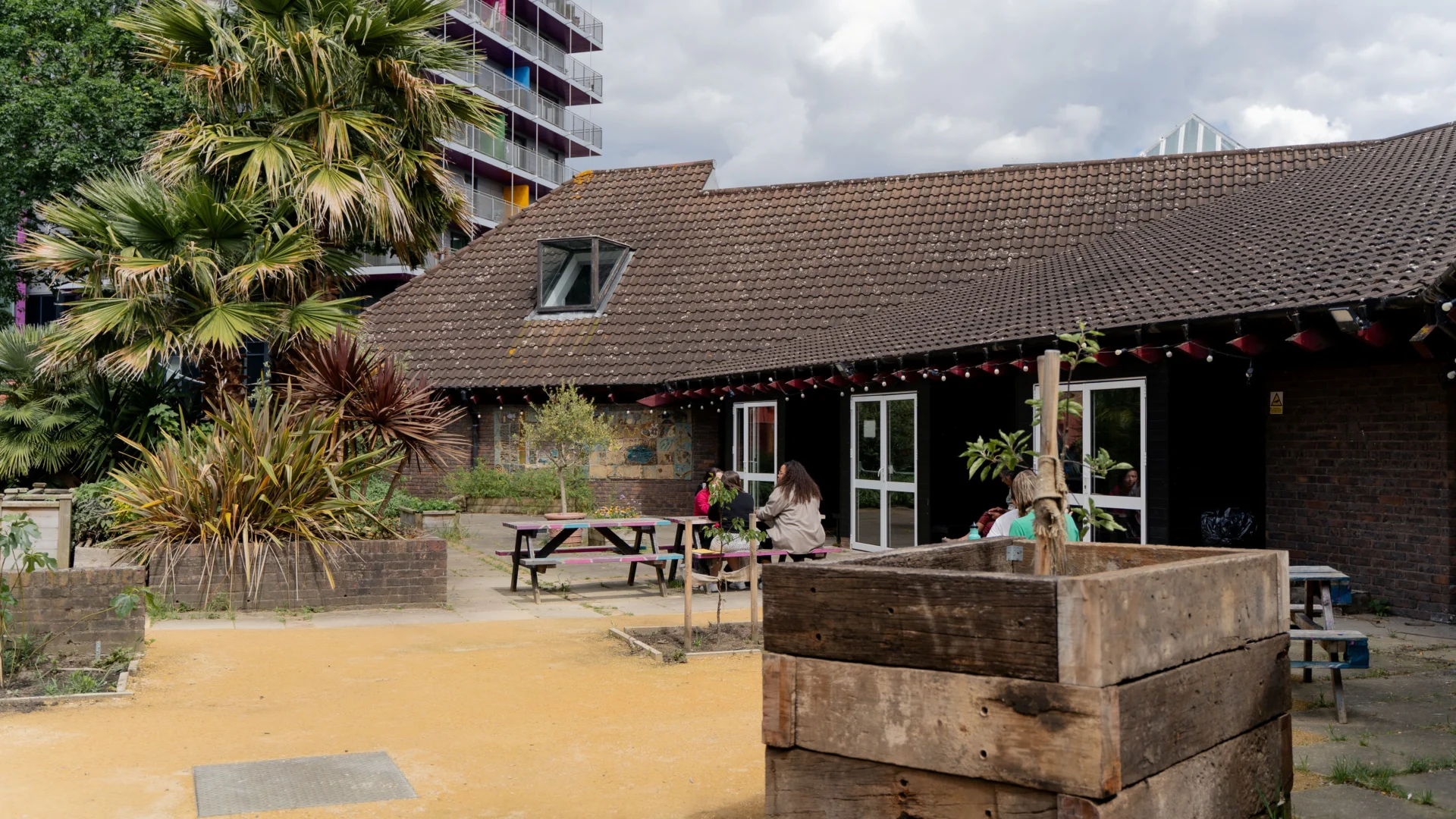 A photo of the Albany garden. A large palm tree is in the centre of the space, with people sat on picnic benches around it. In the foreground of the image is a large wooden planter with a young tree growing in it. At the back of the image is the side of the Albany building - a squat brick building with a tiled roof.