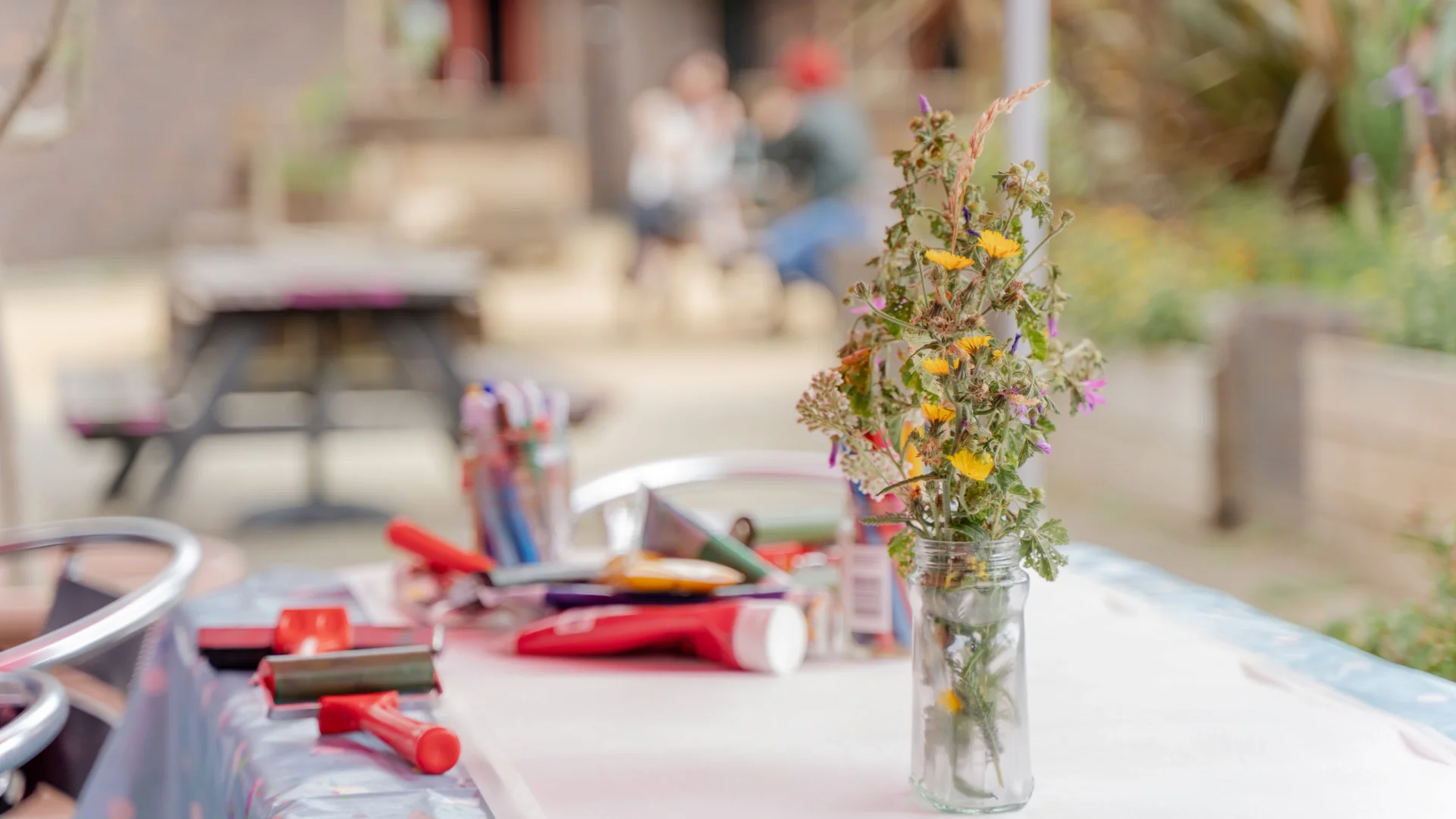 A photo of a table in the Albany garden, with a vase of dried flowers, paper, paints and paint rollers on it.