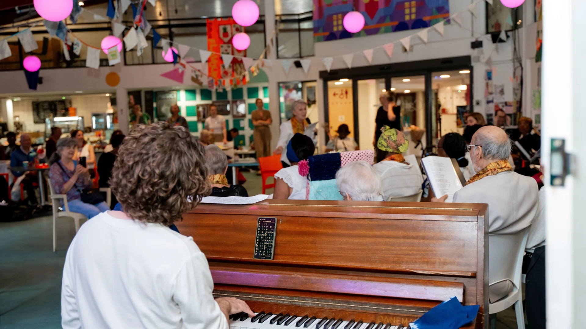 A photo of the Meet Me choir performing in the Albany cafe. The view is from behind the pianist, looking over the piano to the choir and audience beyond.