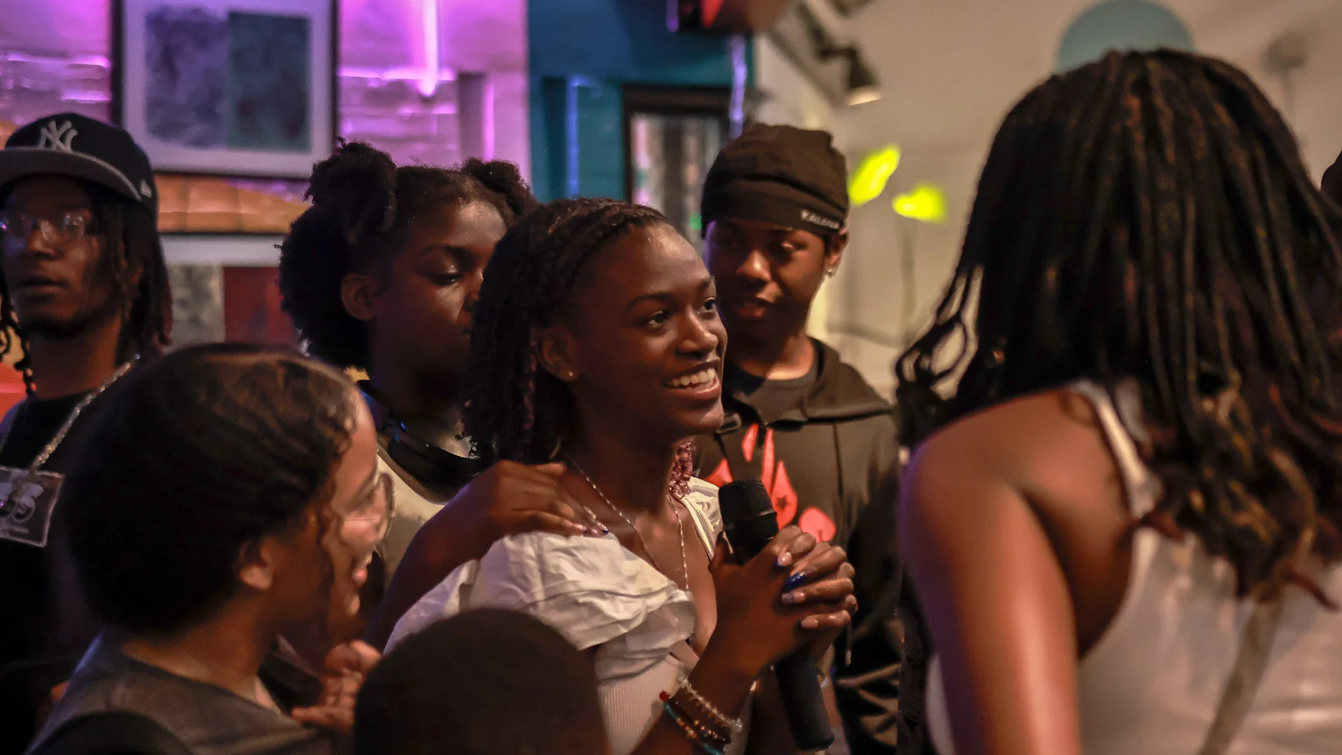 A group of young people gathered in a lively indoor space, with one girl in the centre smiling and holding a microphone.