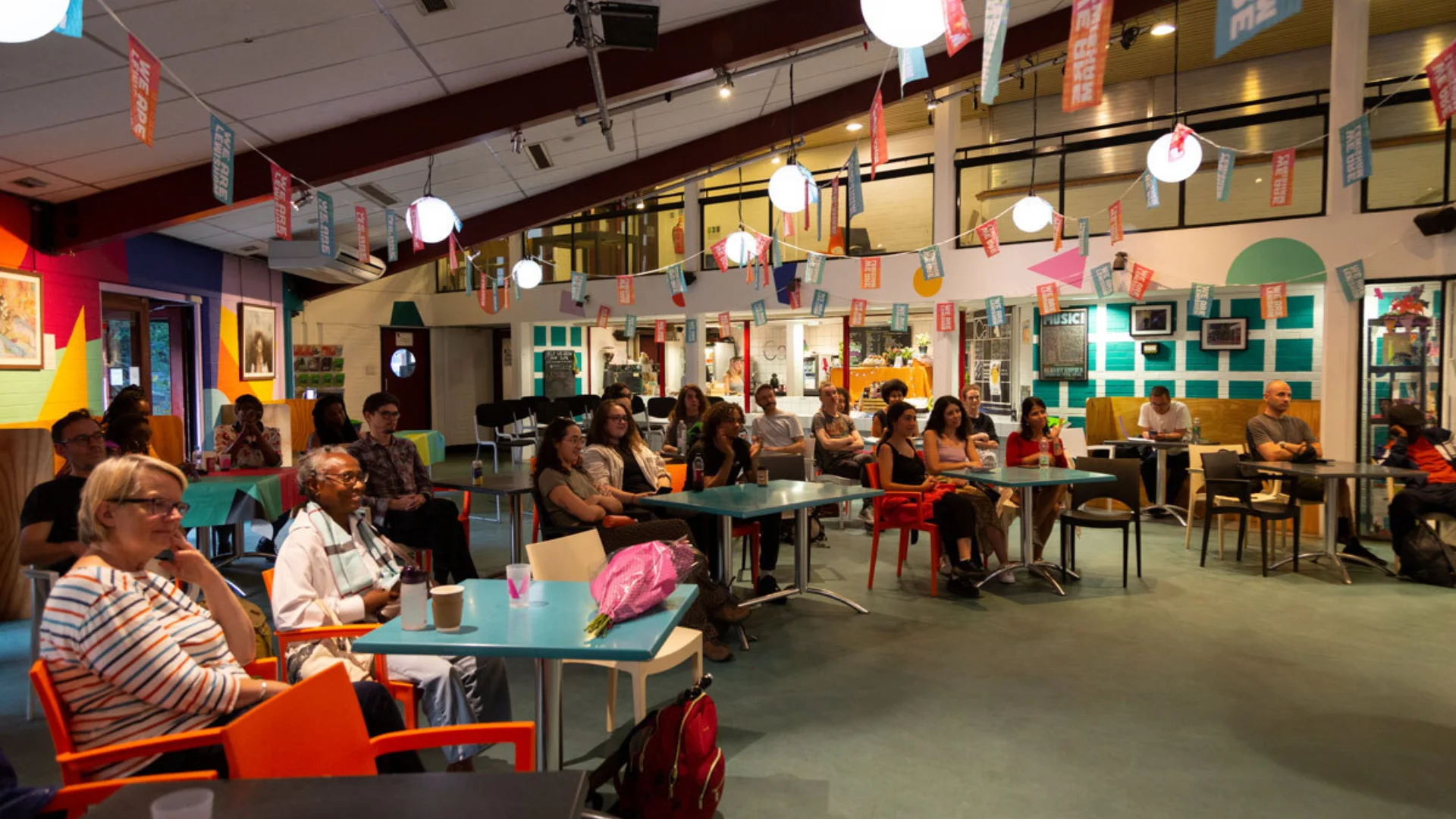 A diverse audience of many ages sat inside a colourful cafe space. All look very engaged and attentive.