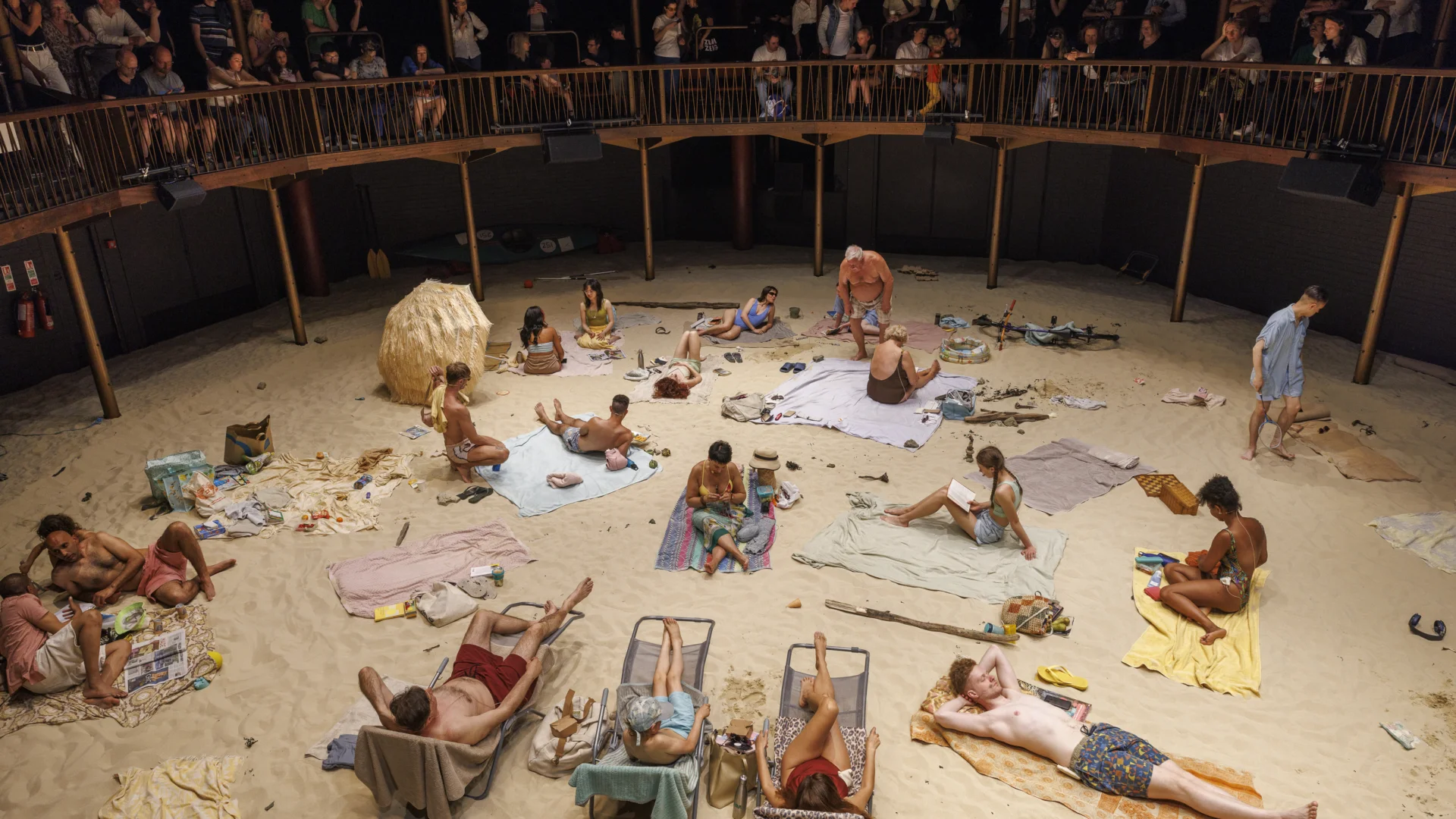 A view looking down at the Albany auditorium from the balcony. The floor is covered in sand to resemble a beach, with performers sunbathing on sun loungers and towels.