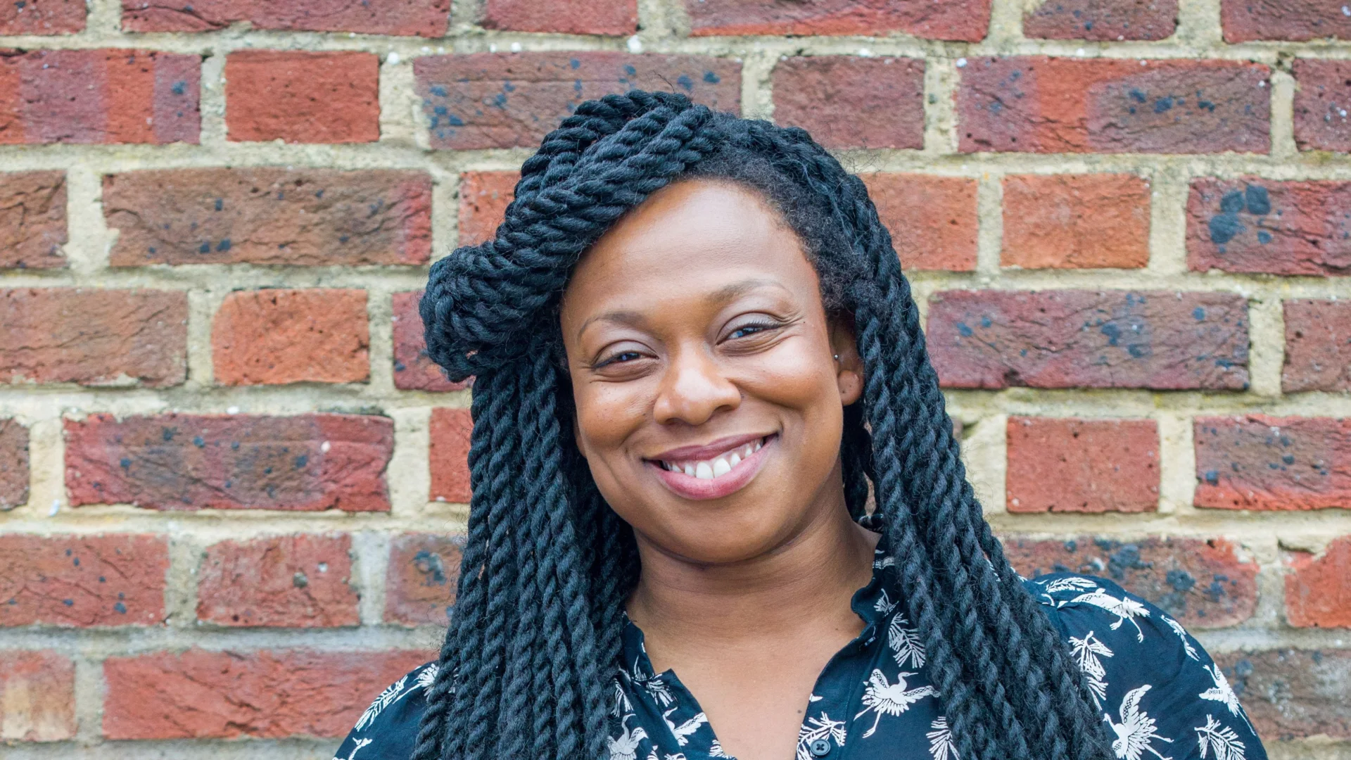 A photo of Vicki Amedume, a black woman with long braids, stood in front of a brick wall.