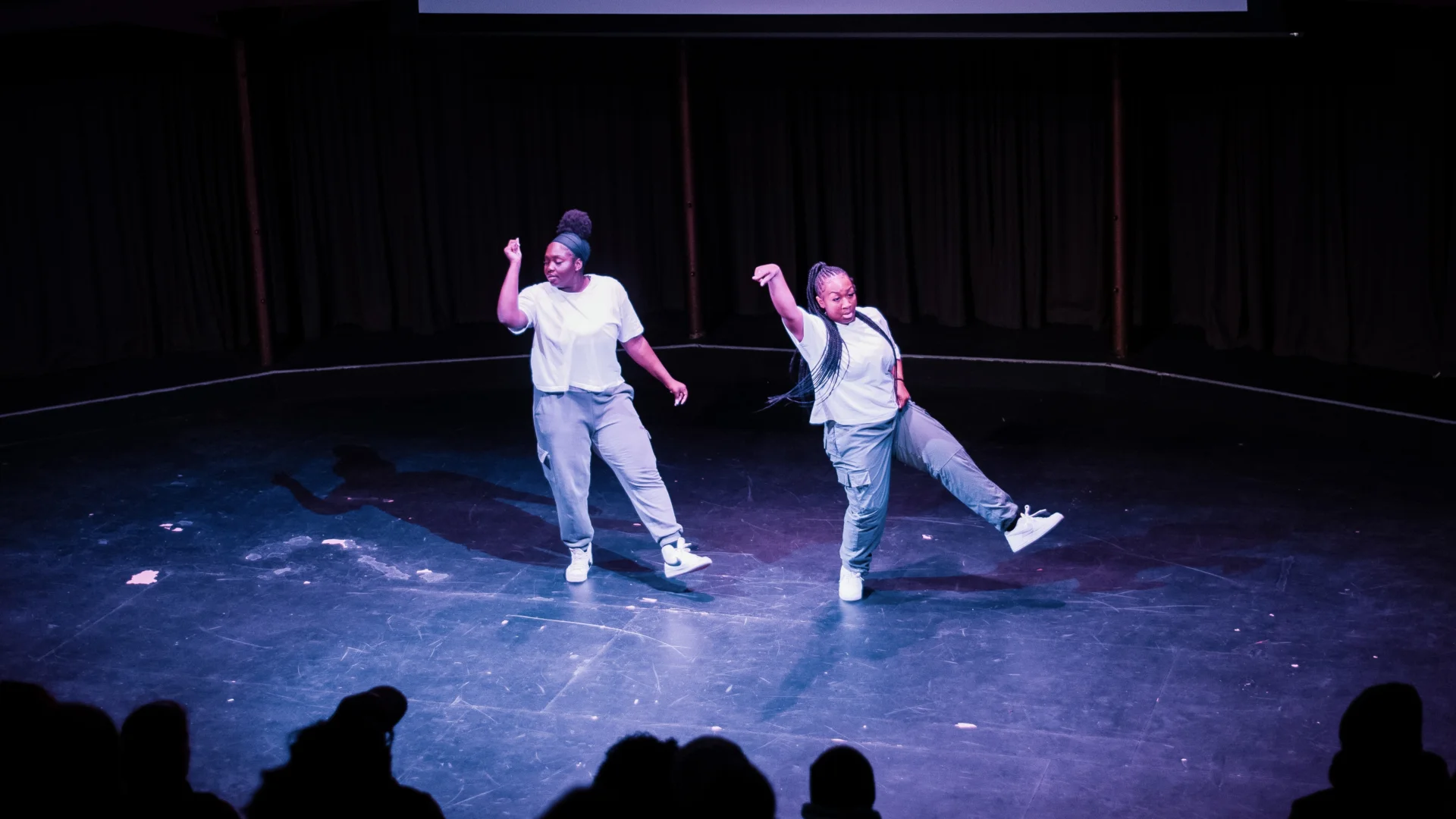 Two young dancers in a stage under spotlight. They are moving in sync and the audience is silhouetted in the foreground of the image.