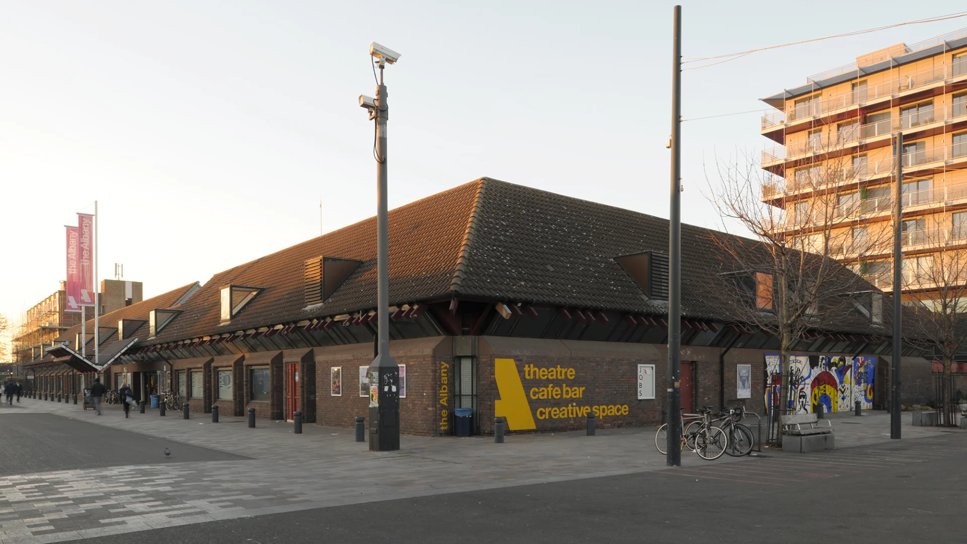 An exterior shot of the Albany building from Deptford Market Yard. It is a squat brick building with pink flags above the entrance. The photo is taken on a cloudy day.