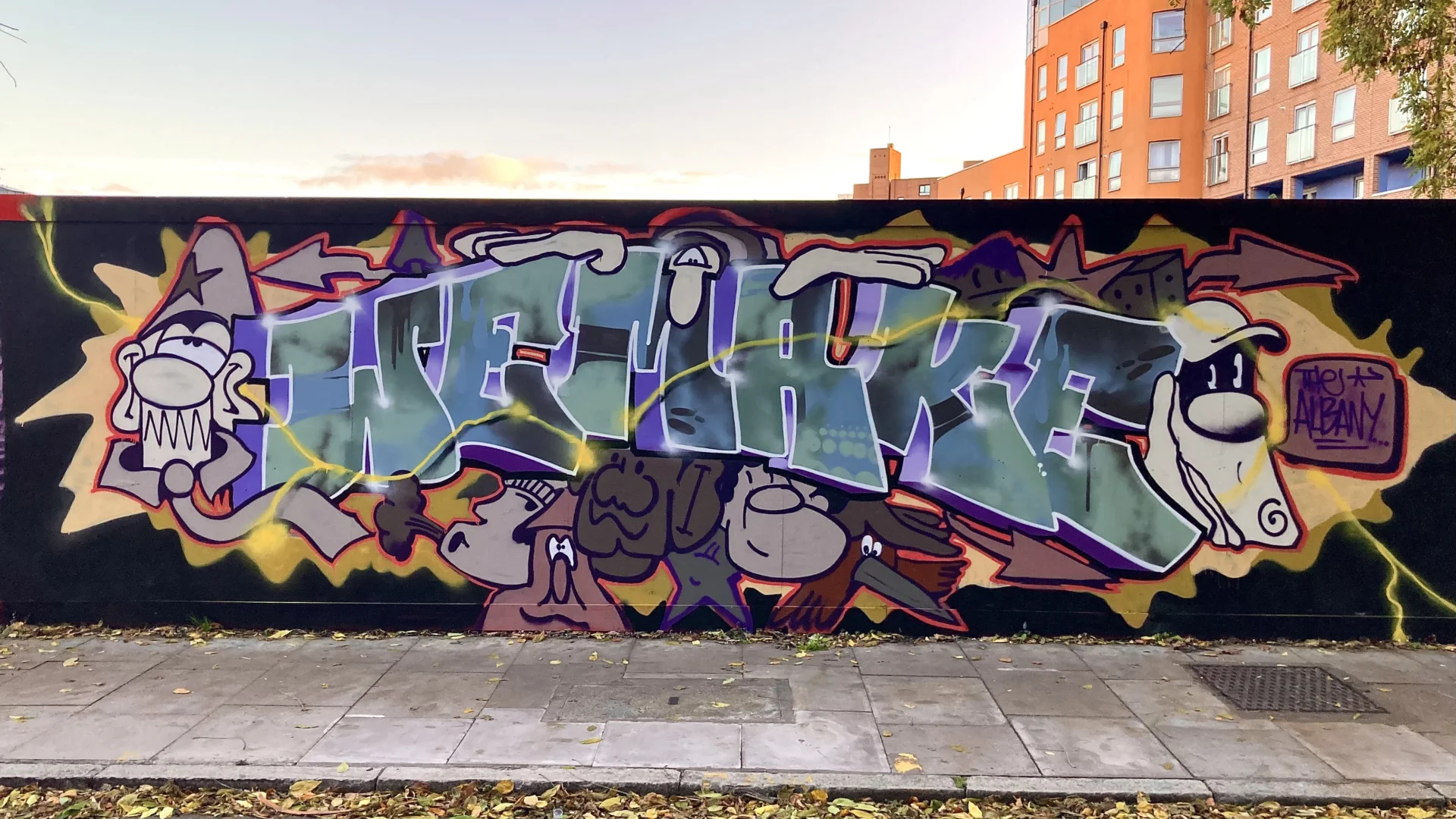 An image of a wall painted black with blue and purple grafitti on it. The grafitti is blue text that says 'We Make' with purple cartoon characters around it.