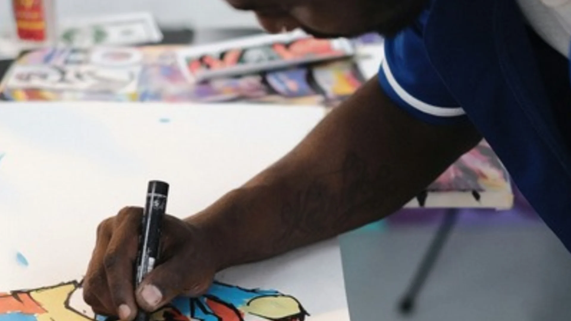 A close up photo of a black man leaning over a table, drawing. He is using a black marker pen to outline an illustration of a face, coloured in with blue, orange and yellow.
