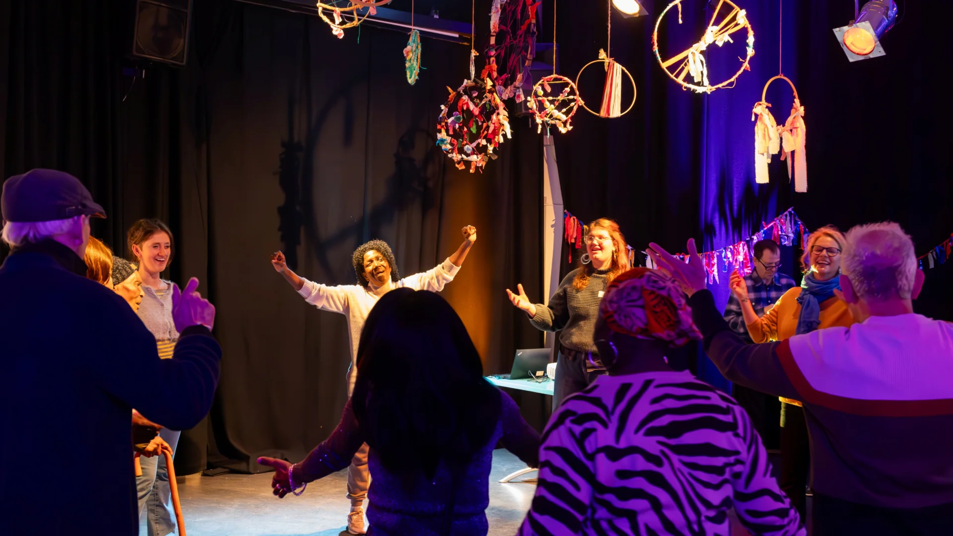 A group of people stand in a circle with their arms raised (they are doing a dance warm up). The room is surrounded with black curtains and there is purple lighting, and dream catchers hanging from the ceiling.