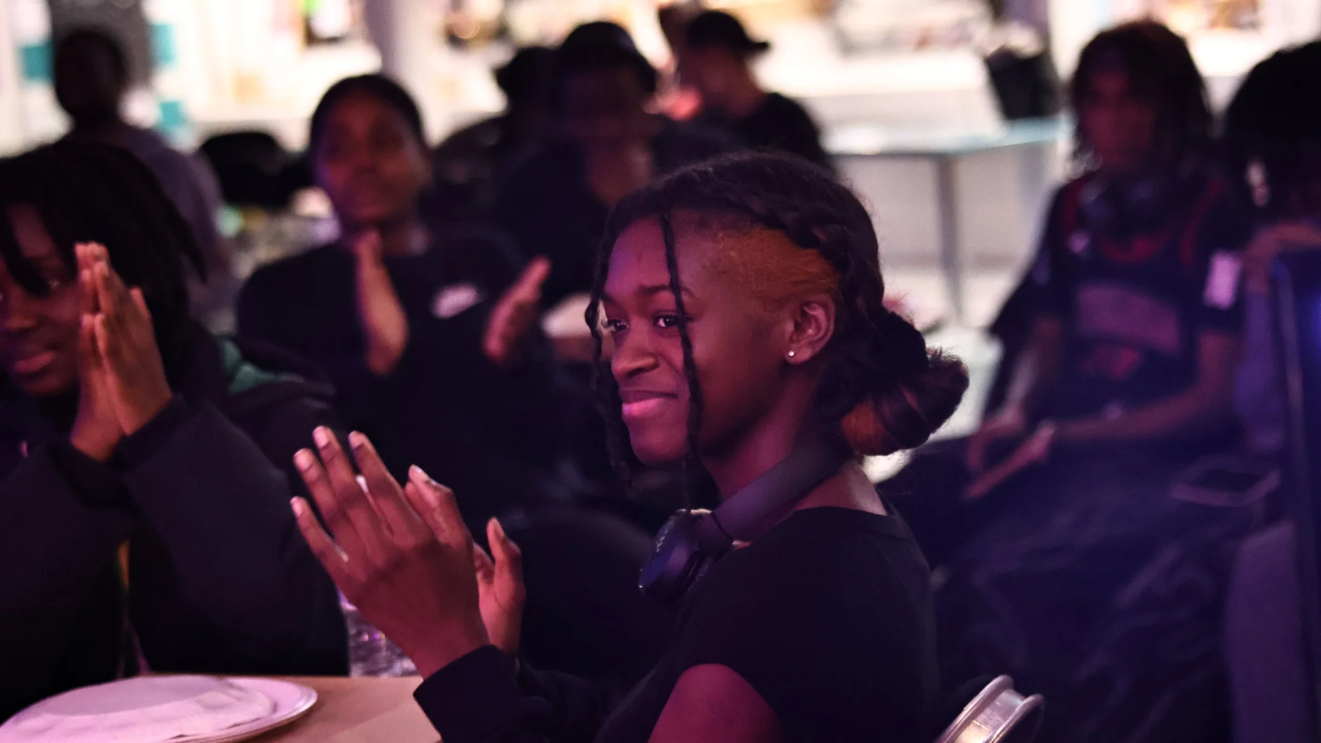 A photo of a young black girls sat in a crowd. She is wearing a black hoodie, looking off to one side and smiling and clapping.