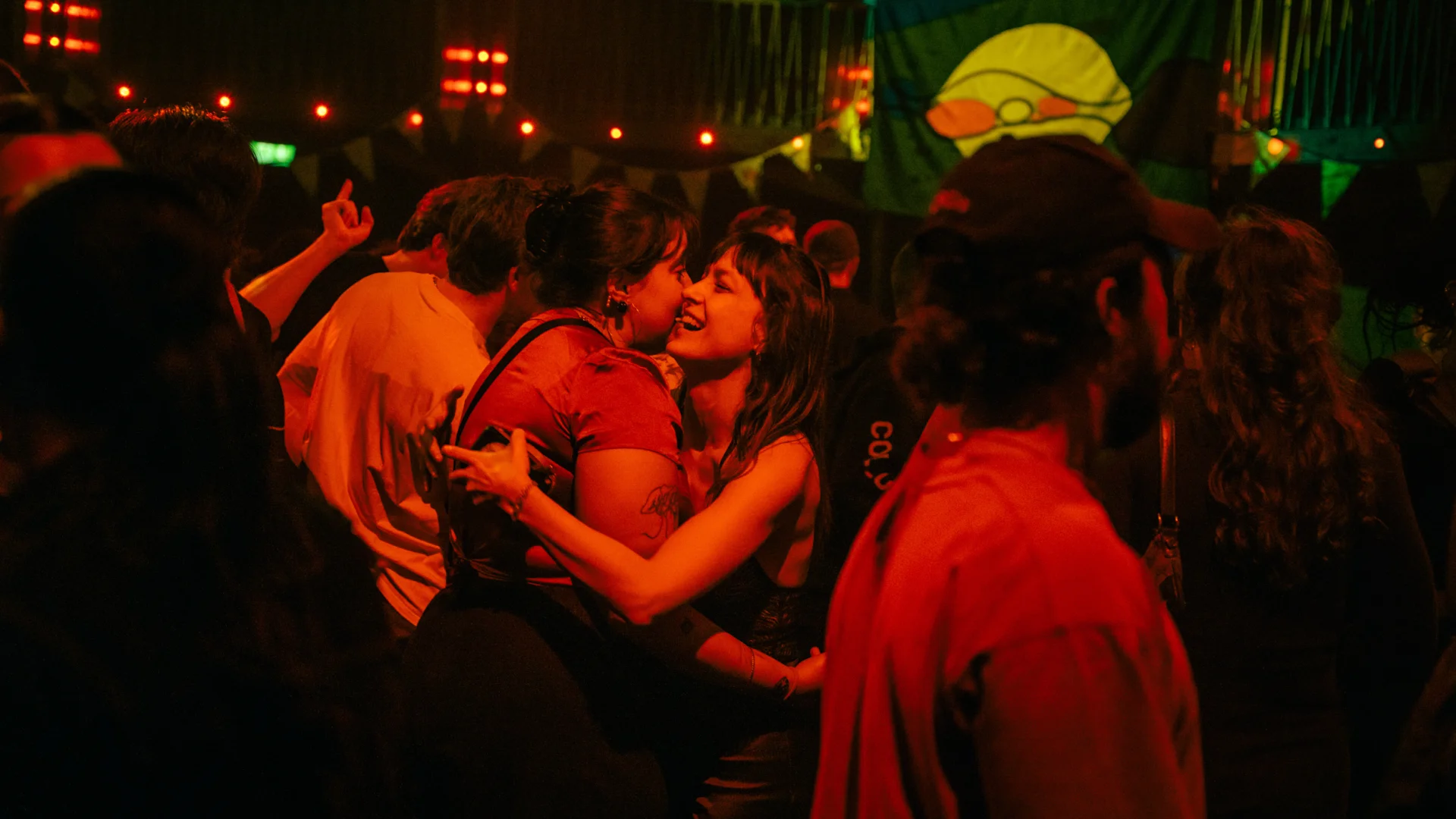 A photo of two women hugging in the middle of a crowded dance floor. They are bathed in red lighting.