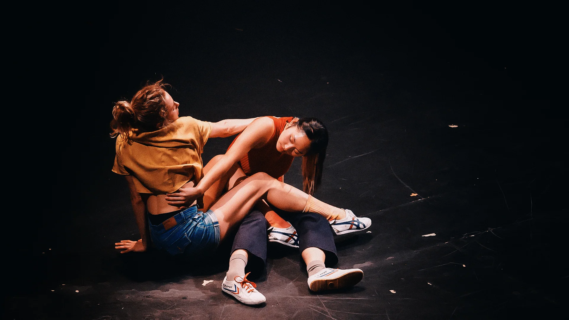 Two female dancers are wrapped around each other on a black stage floor. One is wearing denim shorts and a yellow top and is reaching towards the other's shoulder. The other dancer is wearing blue trousers and a red top.