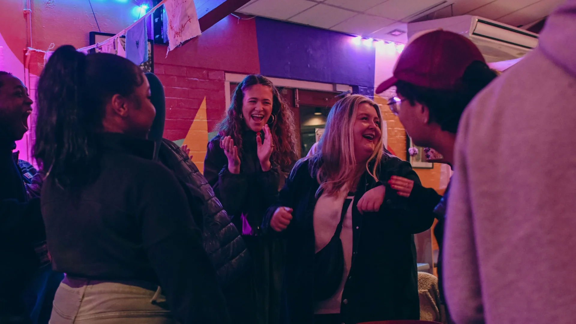 Photo of a group of people chatting and laughing in the Albany cafe. There is a colourful mural in the background and they are bathed in a purple light.
