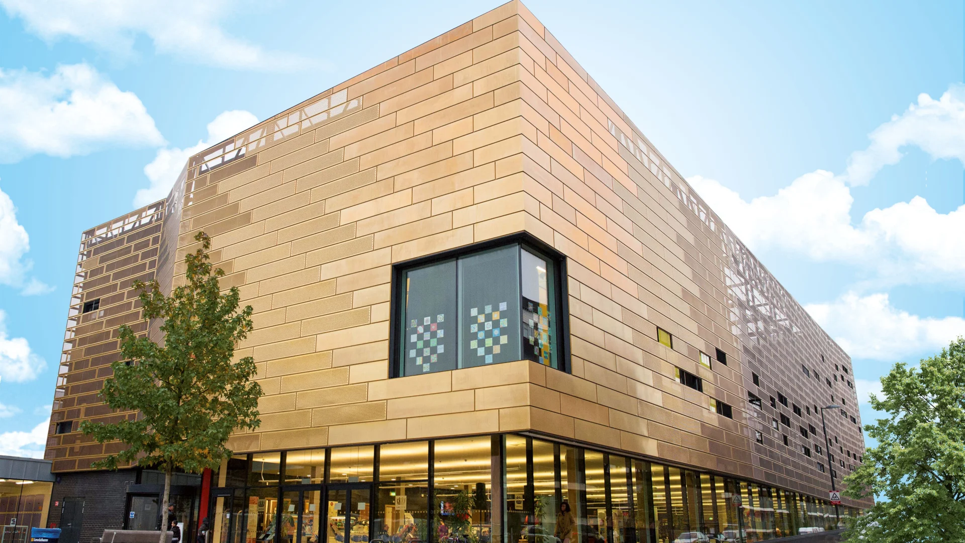 An external shot of Deptford Lounge - a large gold building, with glass windows on the ground floor. It is a summer day and there are white clouds in the sky, and green trees in front of the building.