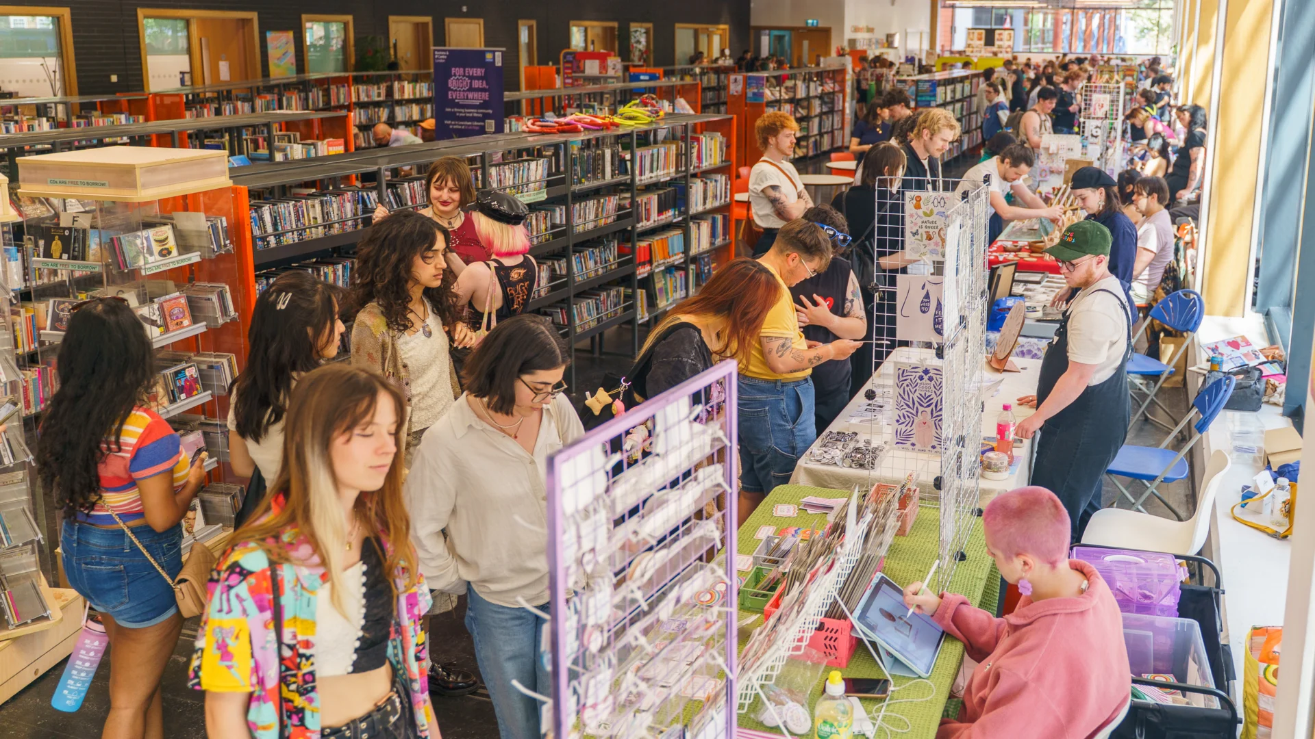 A row of craft stalls in the library at Deptford Lounge. The stalls are parallel to the bookshelves in the library and people are milling around, chatting to the stallholders.
