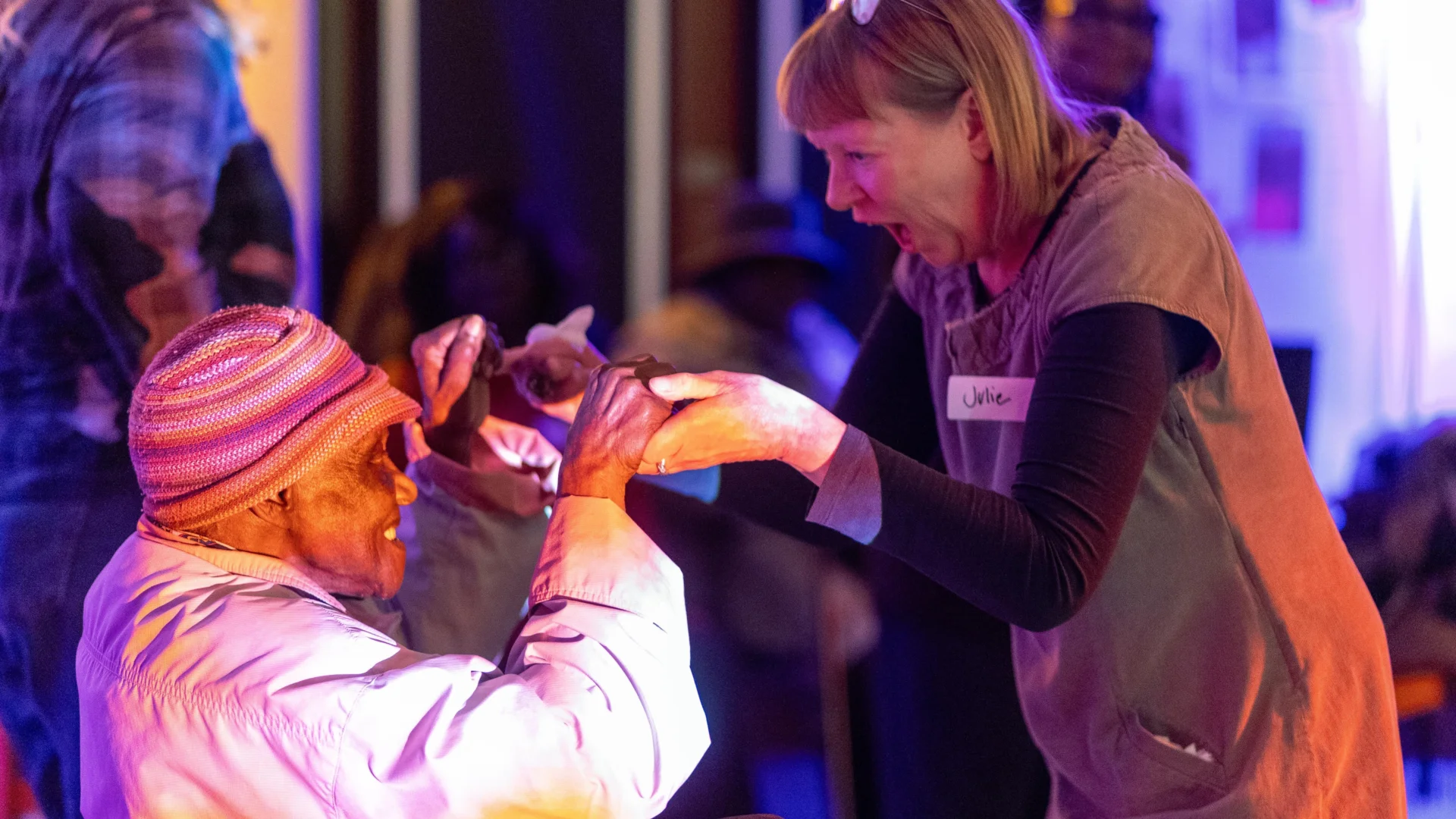 A woman is holding the hands of, and dancing, with another lady in a wheelchair. Both women are smiling enthusiastically at each other giving a joyful and excited.