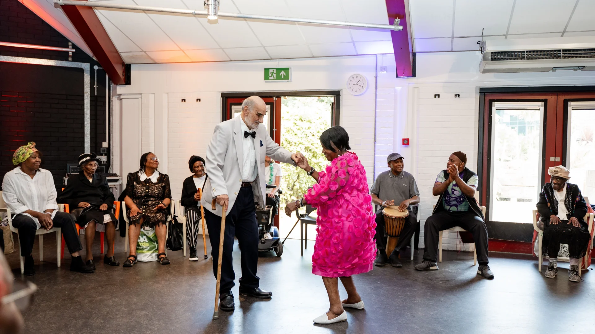 There are two older people, dressed up very smart in a suit and pink dress, they are holding hands and dancing in the middle of a bright room with large windows. There are several other people sat in chairs in a circle around them. These people are smiling, clapping and even one man playing a drum. The atmosphere is joyful and lively.