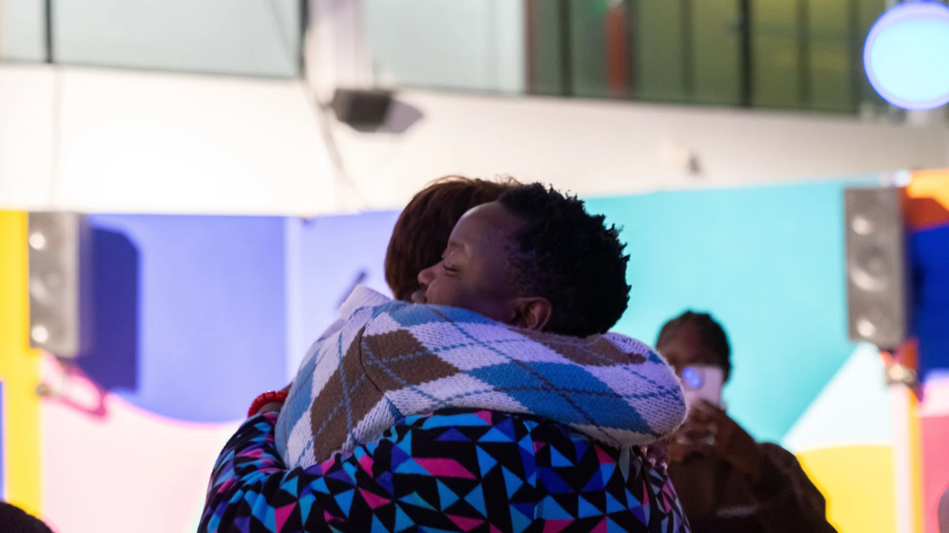 Two people embracing in a warm hug. They are in a colourful, vibrant indoor space with soft lighting.