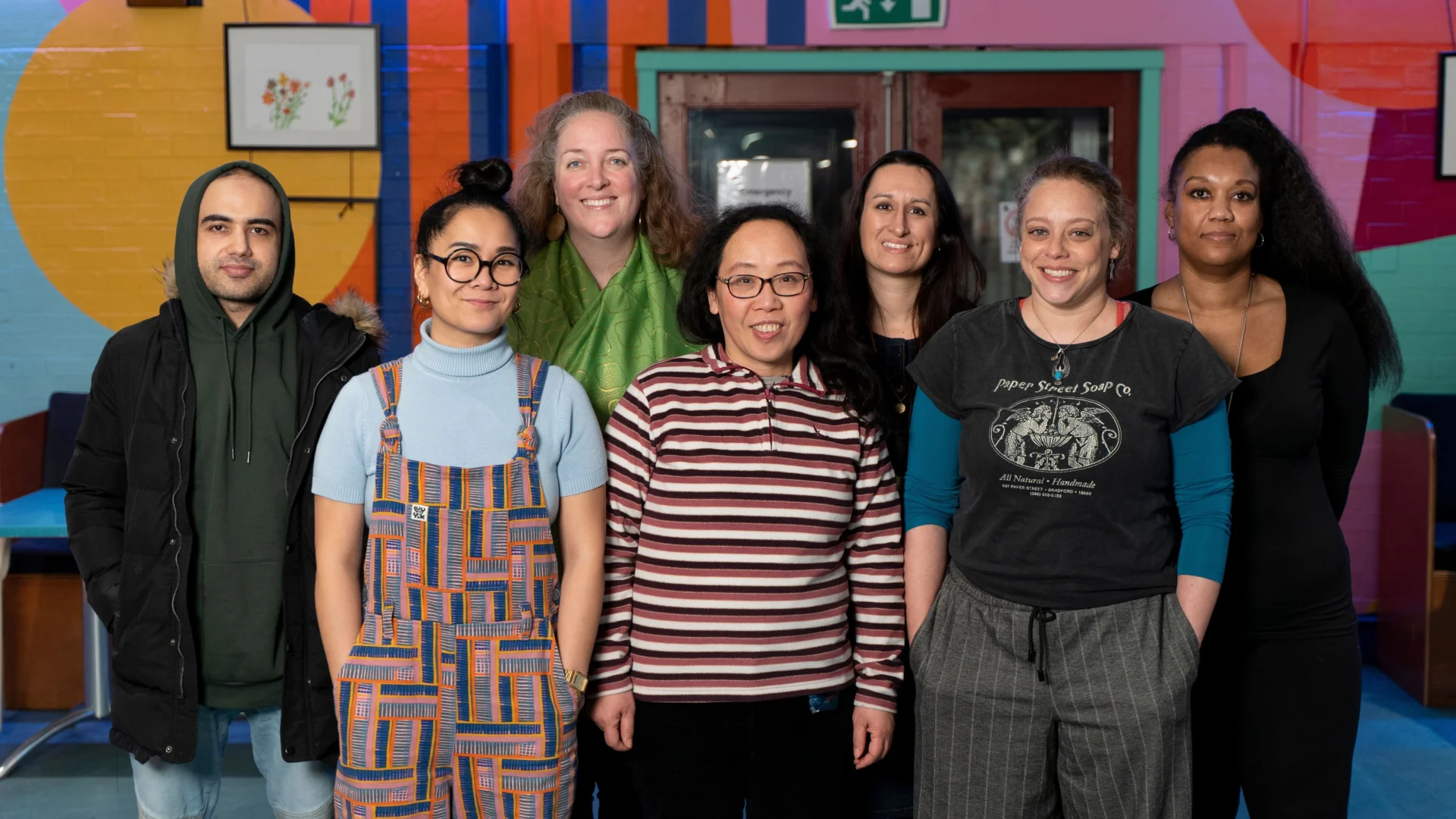 Seven people stand indoors in a row, posing for a group photo against a colorful mural background.