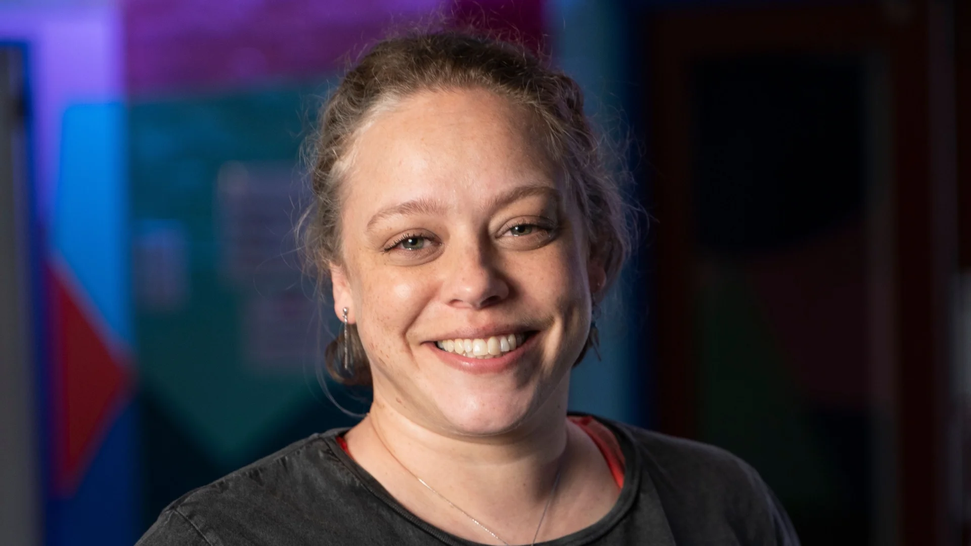 A woman with light hair tied back, wearing a black t-shirt and blue undershirt, smiles in a brightly lit indoor setting with colourful walls.