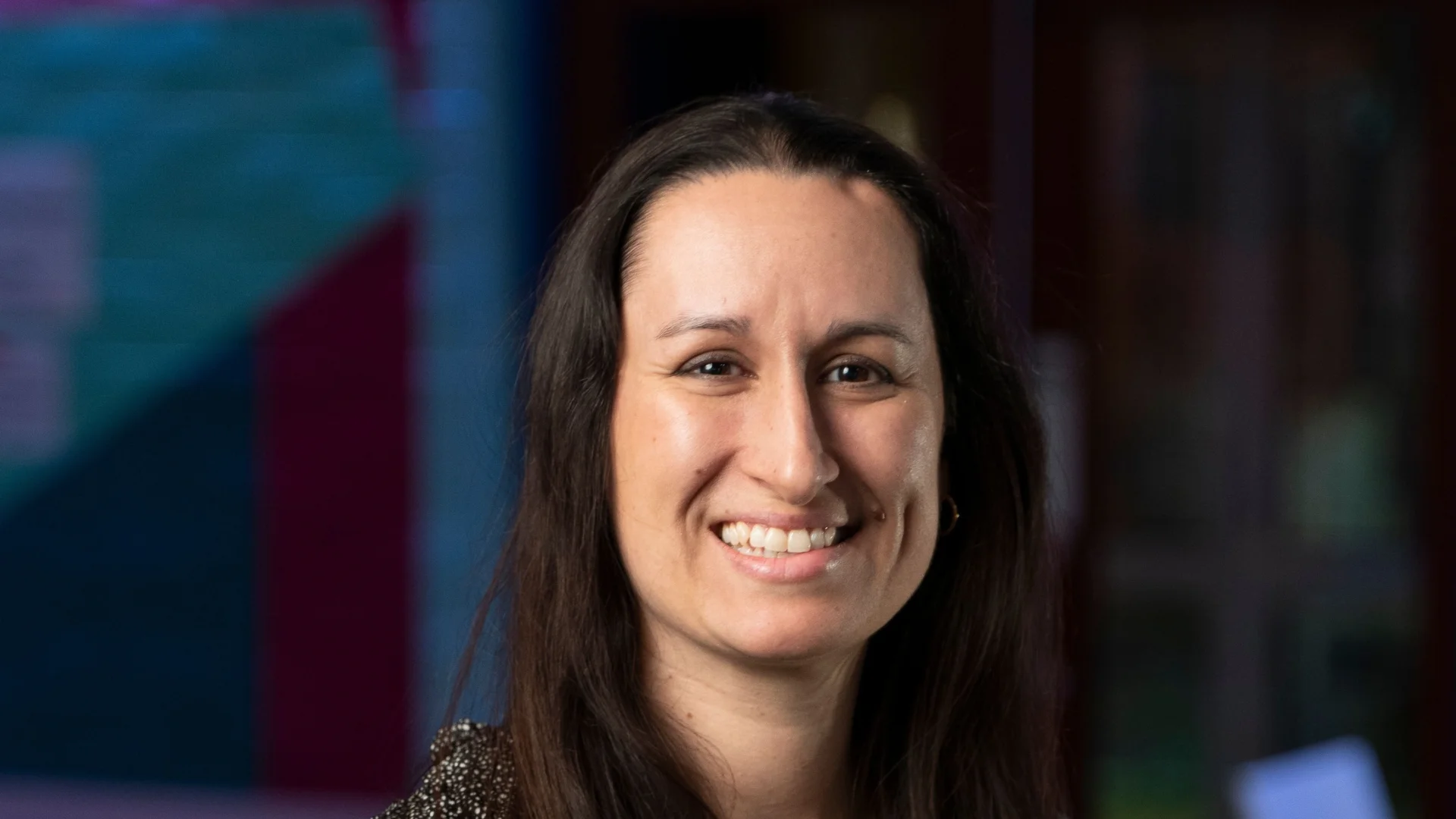 Smiling woman with long dark hair stands in a colourful room. She wears a speckled blouse, conveying a friendly and welcoming demeanor.