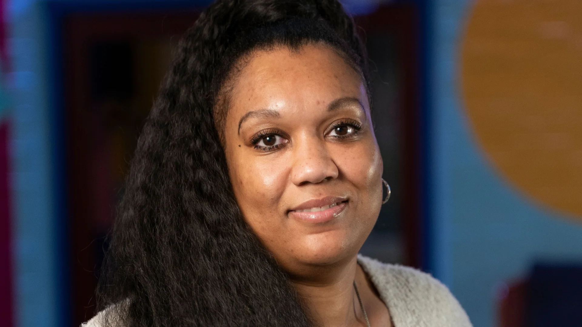Woman smiling in a colourful room with bright lighting. She wears a beige sweater and long necklace, creating a warm, welcoming atmosphere.