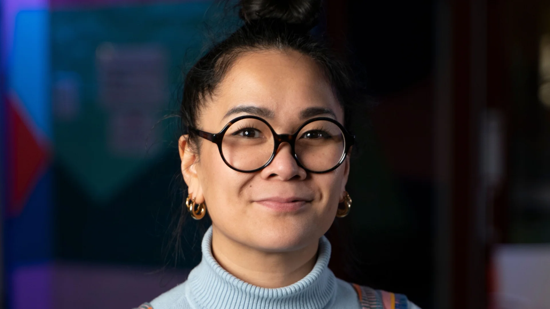 A person with glasses and a hair in a high bun smiles gently, wearing colourful dungarees and a light blue turtleneck. The background is a vibrant, softly lit room.