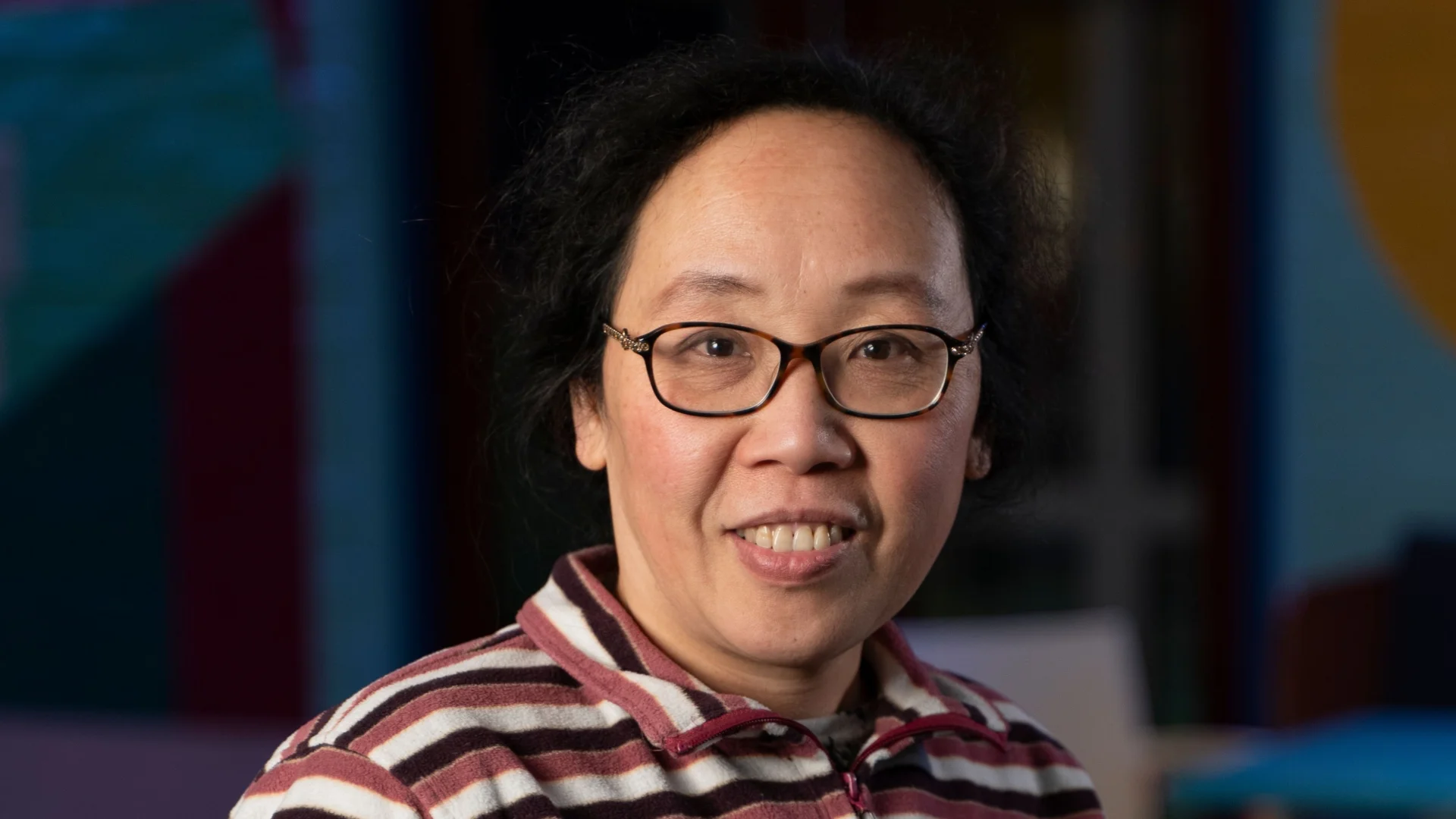 A person in glasses smiles warmly, wearing a striped jumper. The background is colourful, featuring soft lighting, creating a welcoming atmosphere.
