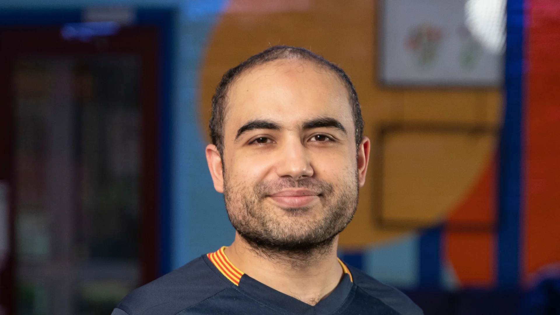 Man smiling, wearing a Barcelona football shirt in a colourful room with vibrant walls. The atmosphere is casual and cheerful.