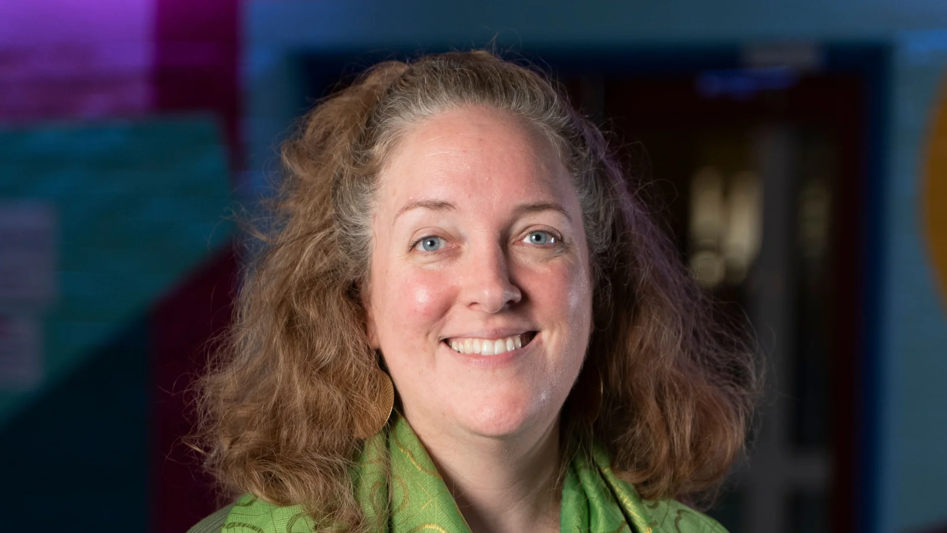 Smiling woman in a green scarf, standing indoors against a colourful background. The atmosphere is warm and welcoming.
