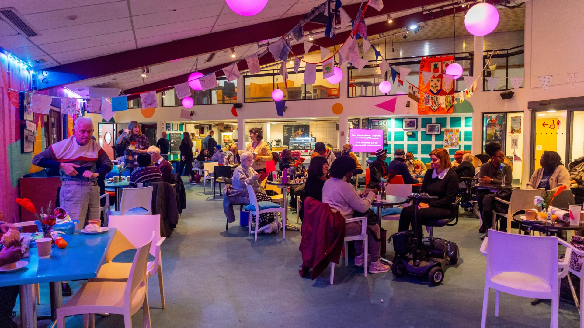 A photo of the Albany cafe during a Meet Me party. The tables are full of people chatting, there is bunting hanging from the ceiling and pink lighting.