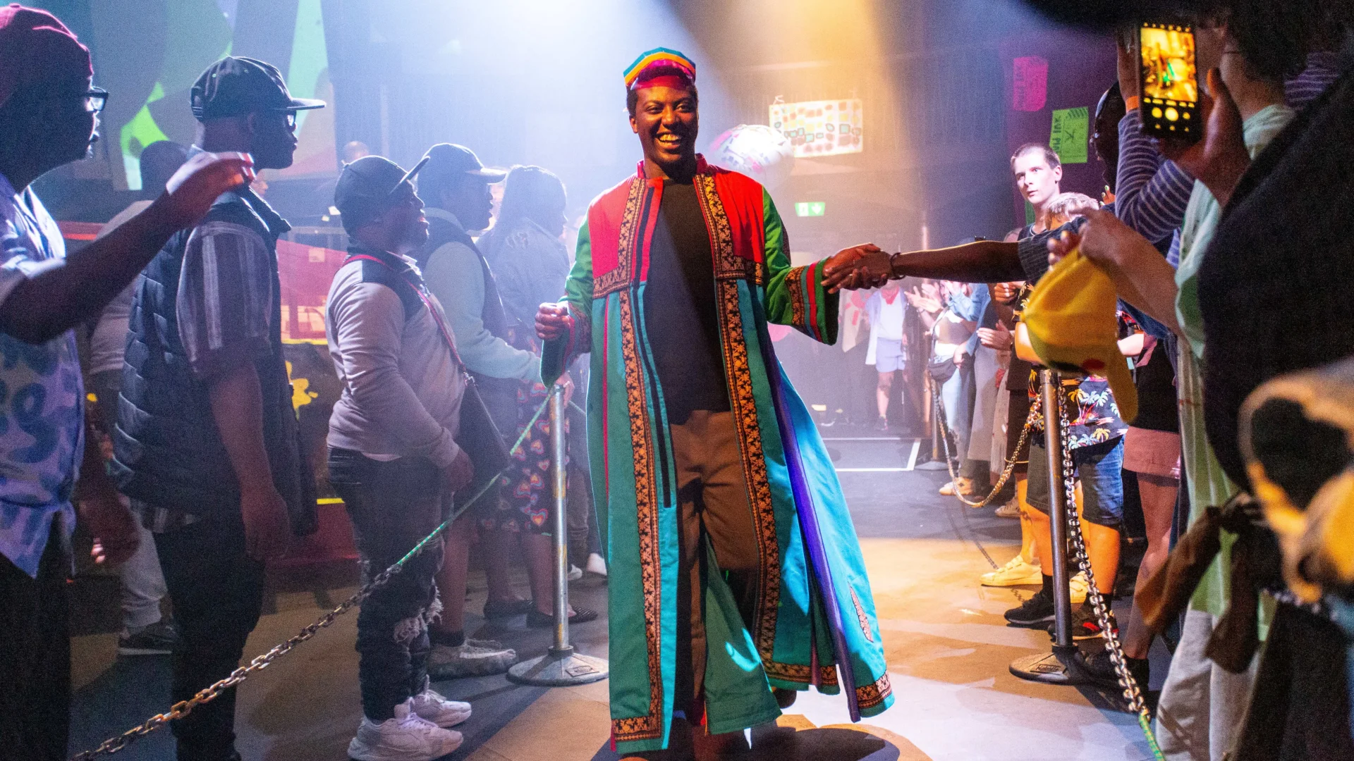 A photo of a young black man wearing bright colours on a catwalk. There is brightly coloured lighting and people on either side of the catwalk are cheering him on.