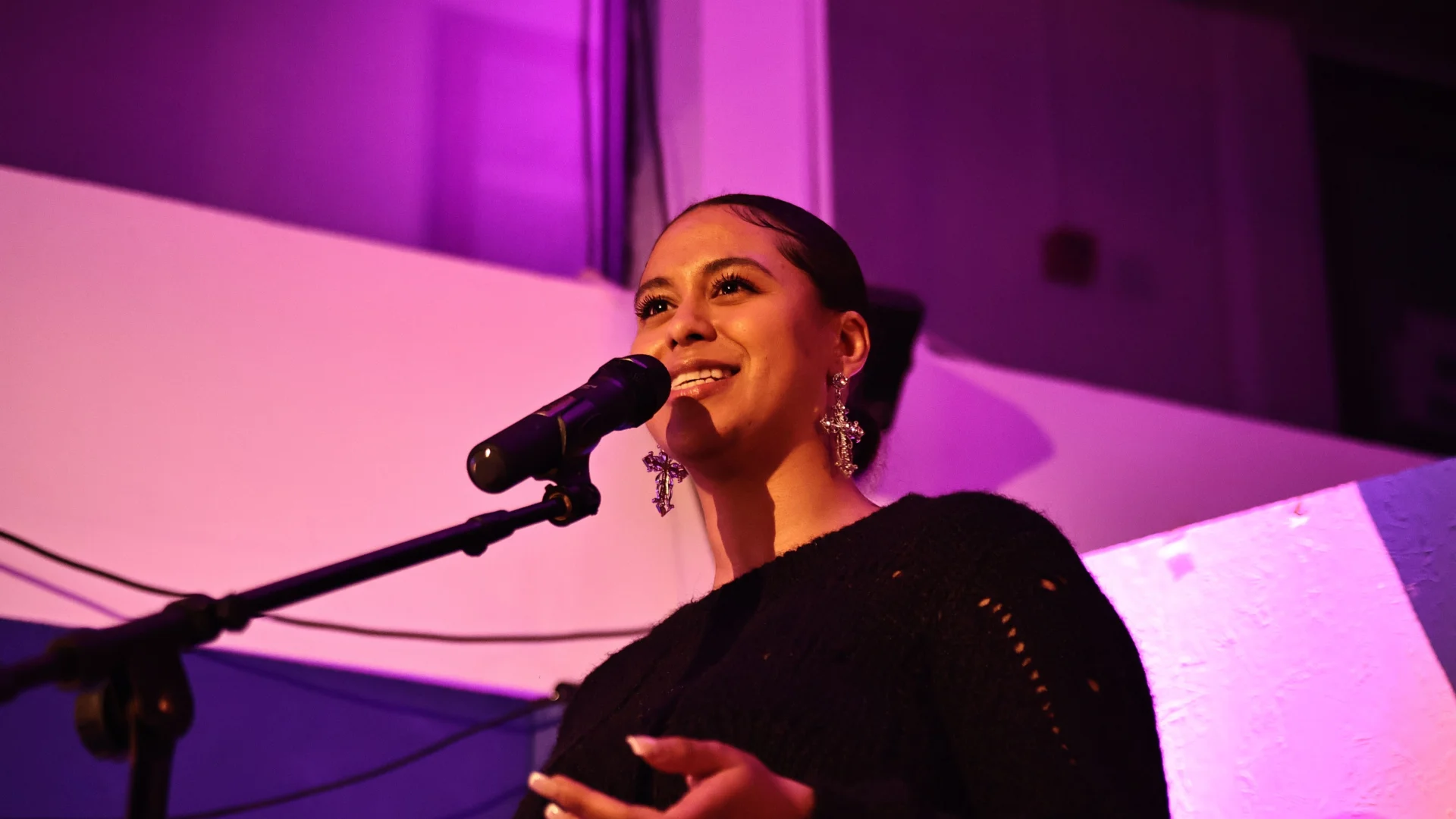 A young woman standing in front of a microphone on stage. The lights are a pink hue and the scene captures her focus she performs indoors, with a smiley demeanor.
