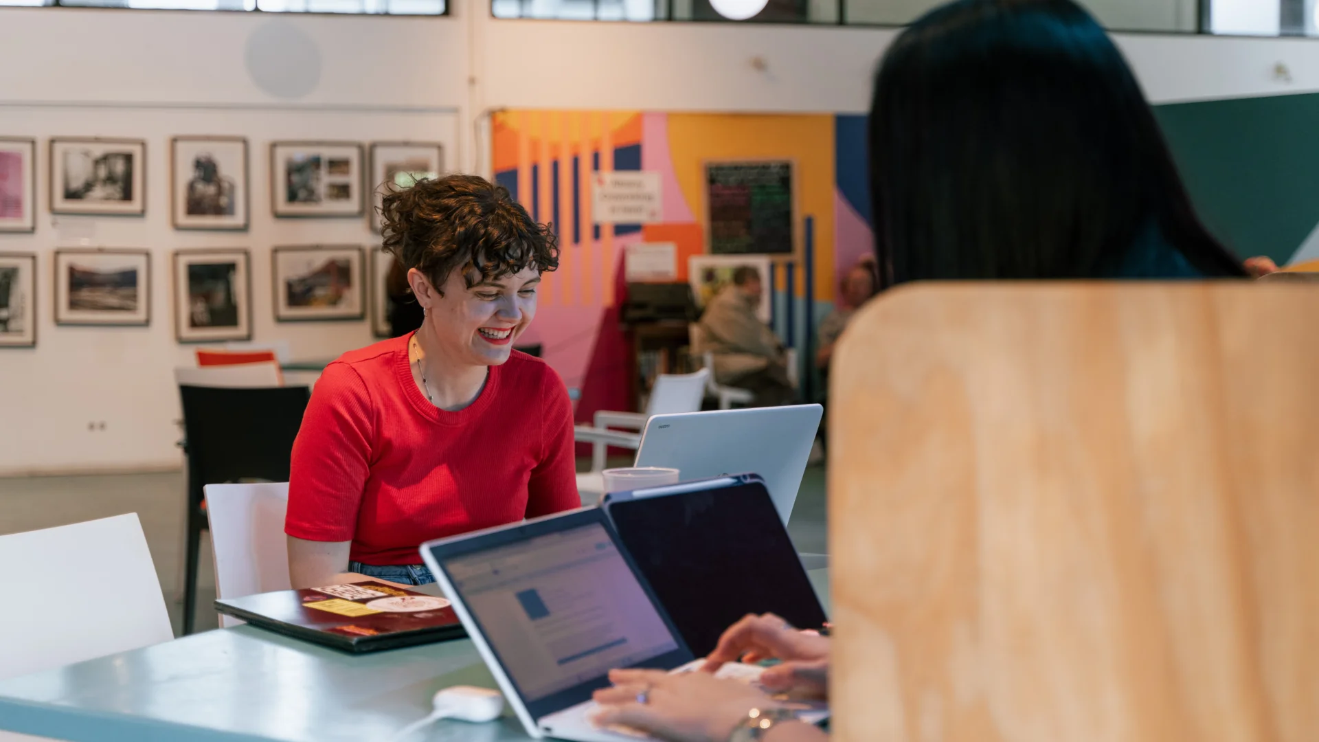 A photo of two people sat at a table on laptops. One has their back to the camera. The other is a white woman with a red top and curly hair, she is smiling.