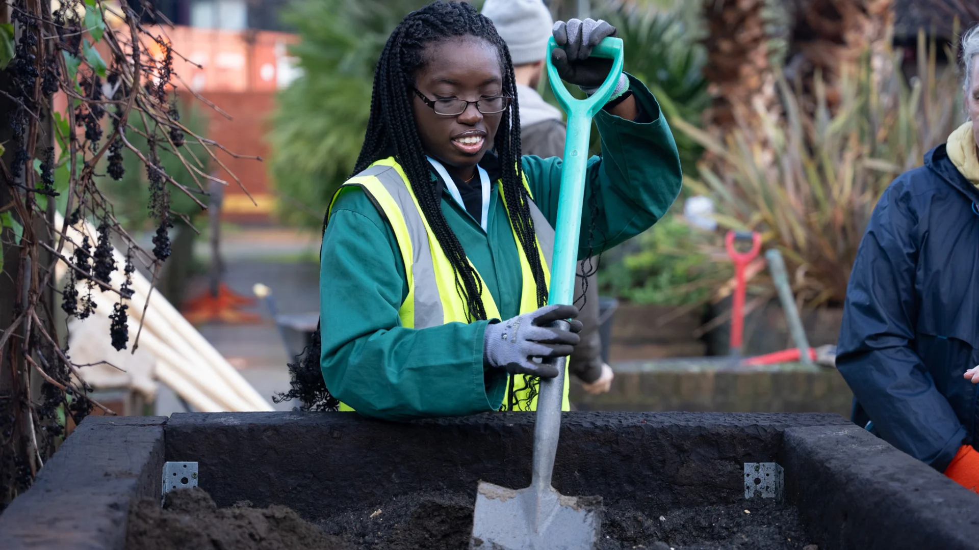 A young girl is holding a shovel in the action of planting a tree in a free standing planter outside. She is wearing a hi-vis vest and smiling.