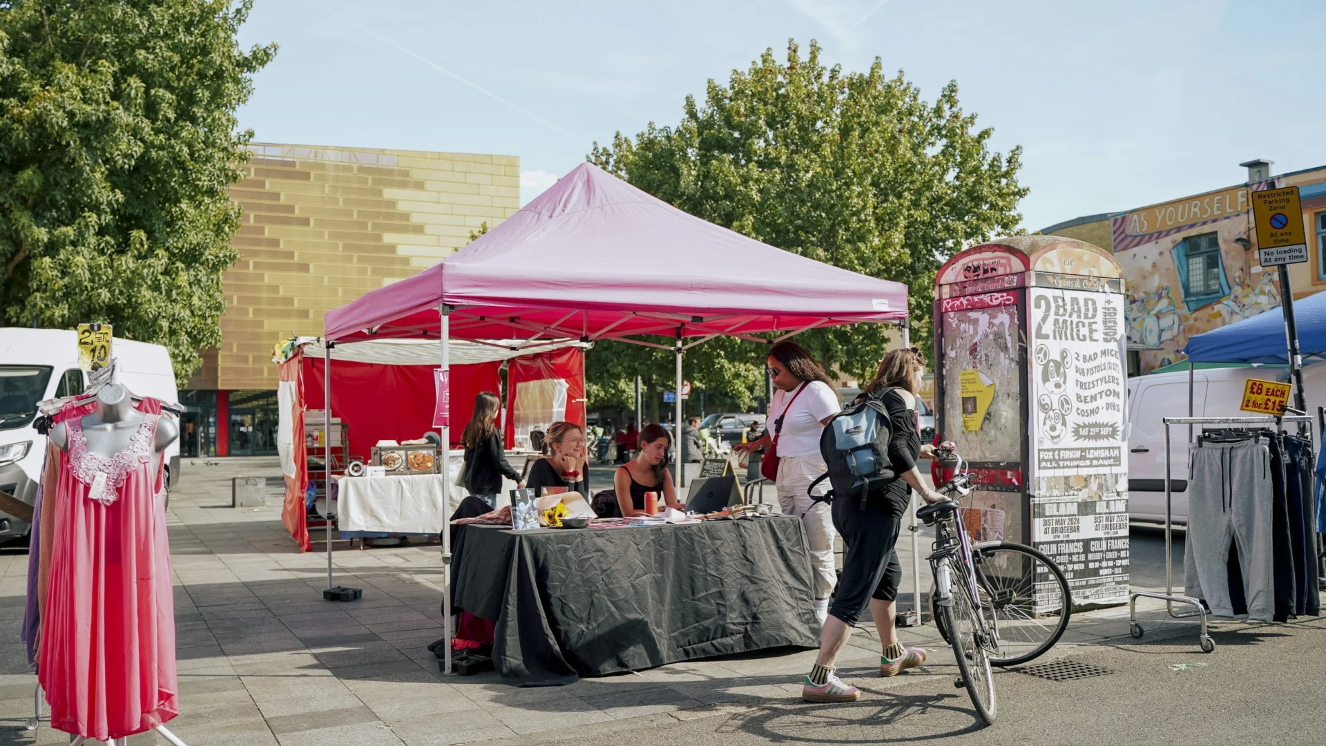 A photo of a stall on Deptford Market. It is a sunny day and the stall is under a bright pink gazebo. Someone holding a bike is stopped in front of the stall and there are trees and other stalls in the background.