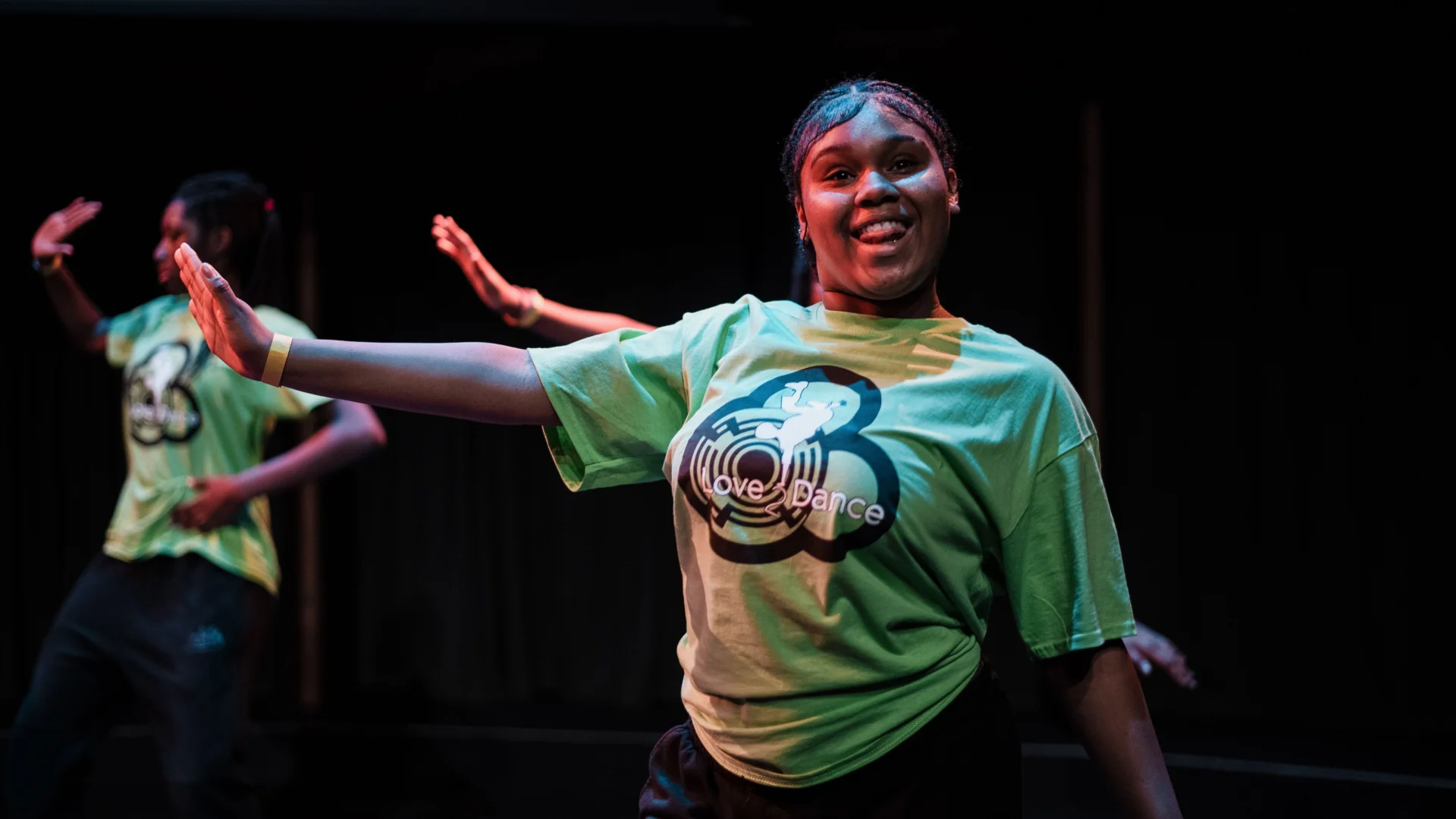 A young woman energetically dances on stage as part of an ensemble, all are wearing a green "Love2Dance" shirt. She's smiling, with arms outstretched, conveying joy and enthusiasm.