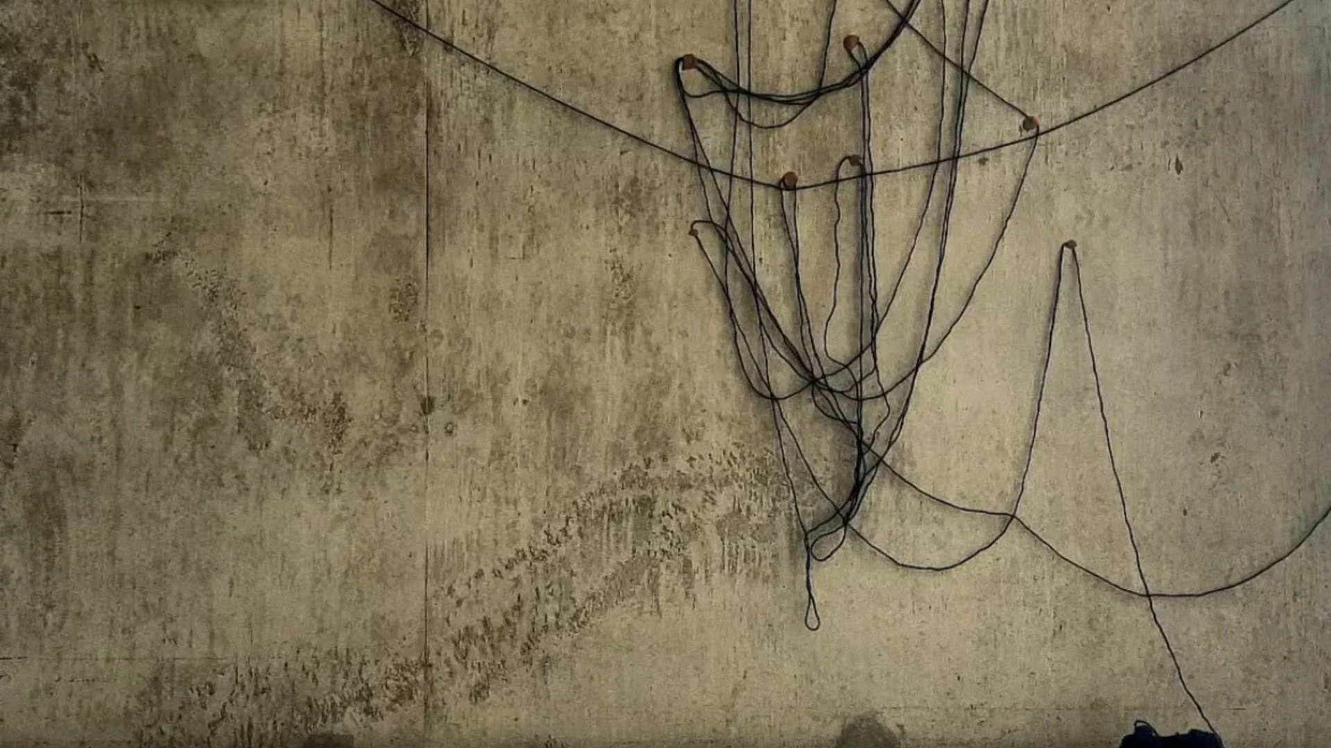 A photo of an artwork. Some black string is hung up on a grey concrete wall and there are papers spread out on the floor.