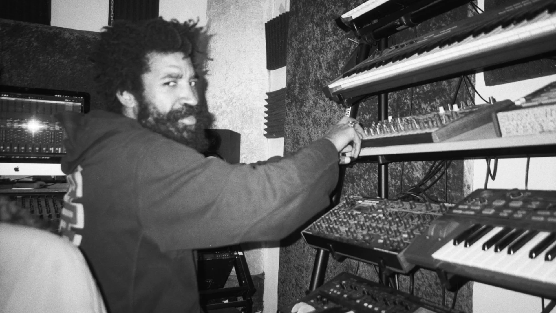 A black and white photo of musician Kwake Bass - a man with an afro, beard and wearing a hoodie, stood in front of a wall of keyboards and DJ decks.