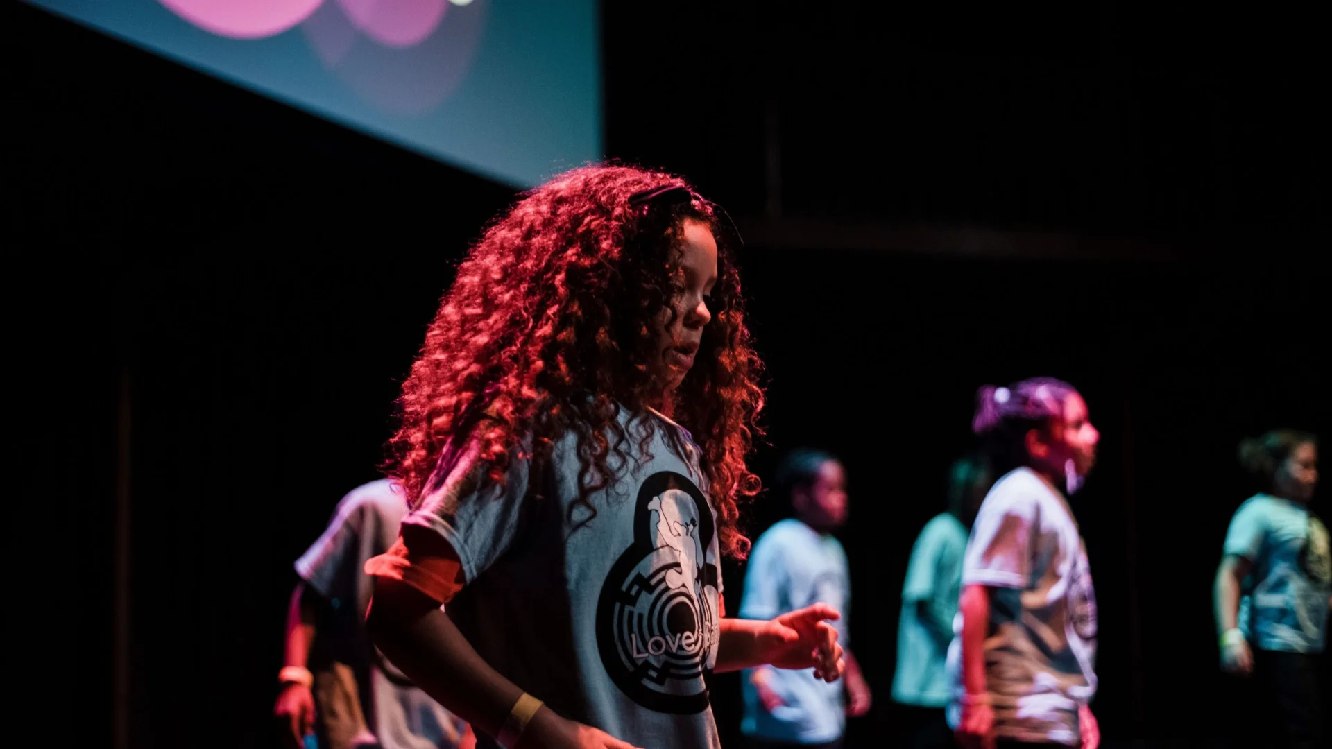 A young girl with long brown curly hair and wearing a blue t-shirt is dancing on stage. Behind her are a group of other children all wearing blue t-shirts.