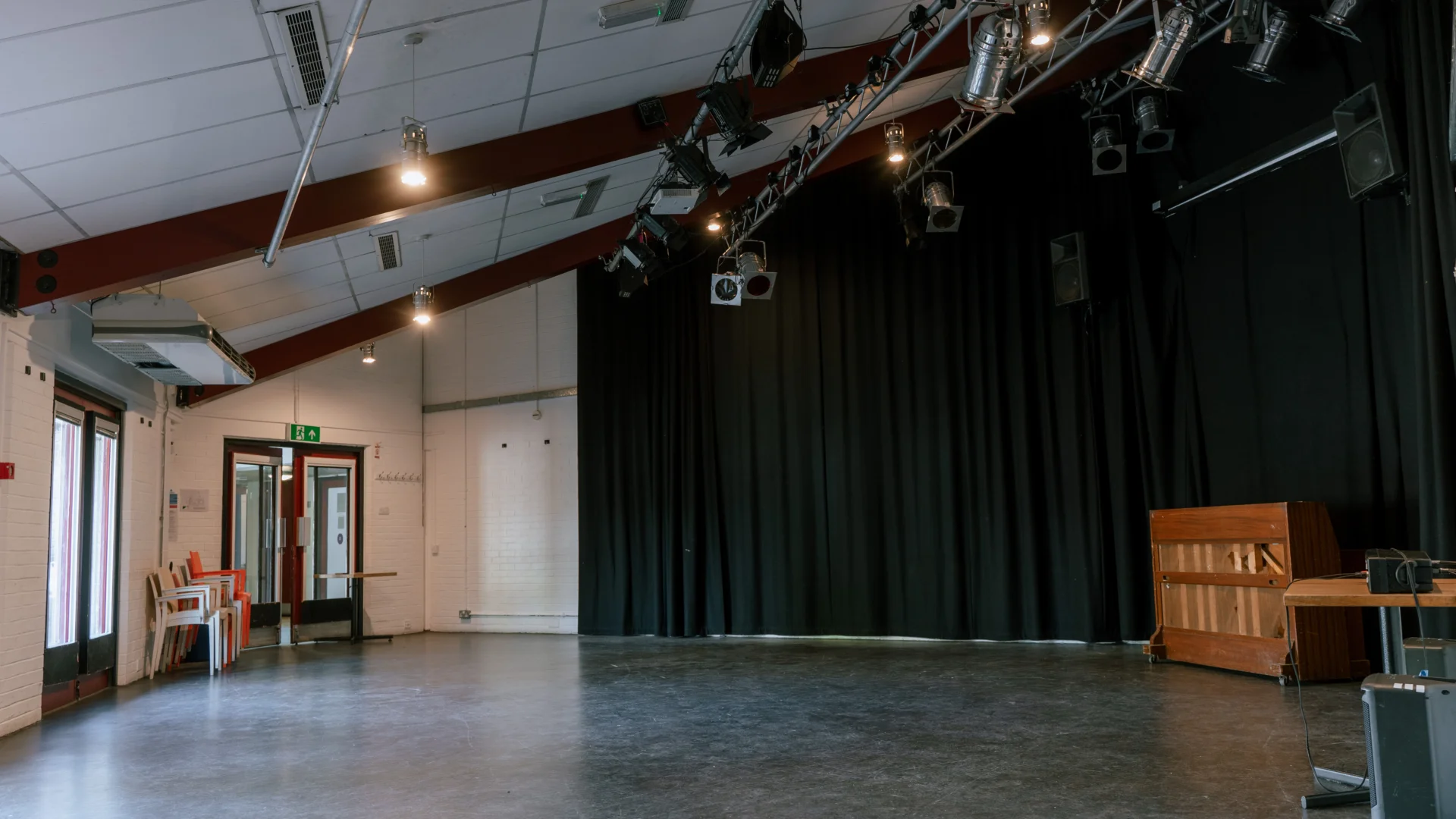 A photo of the Red Room at the Albany. It's an empty space with a lighting rig on the ceiling, and black curtains surrounding the walls. There is a piano in the corner.